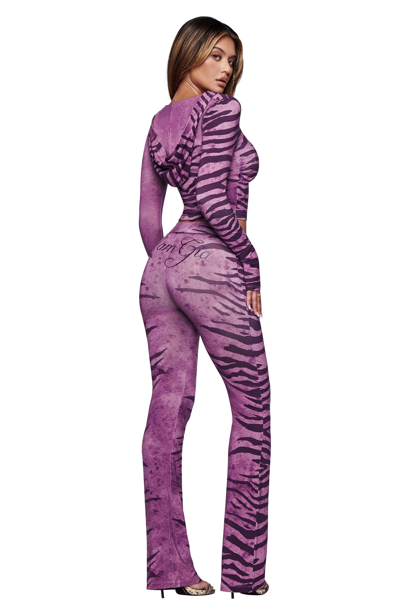 A woman with long, straight hair and high heels faces backward, looking over her shoulder in the I.AM.GIA BLARE HOODIE – PLUM WILD SIDE, a purple hoodie with black zebra stripes, against a white background.