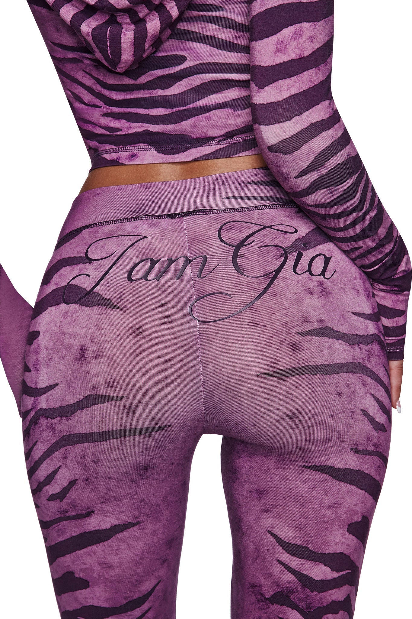 A person wears the I.AM.GIA BLARE TRACKPANT in Plum Wild Side, featuring a flared leg and "I.AM.GIA" script across the back. The image highlights the lower back and upper legs.