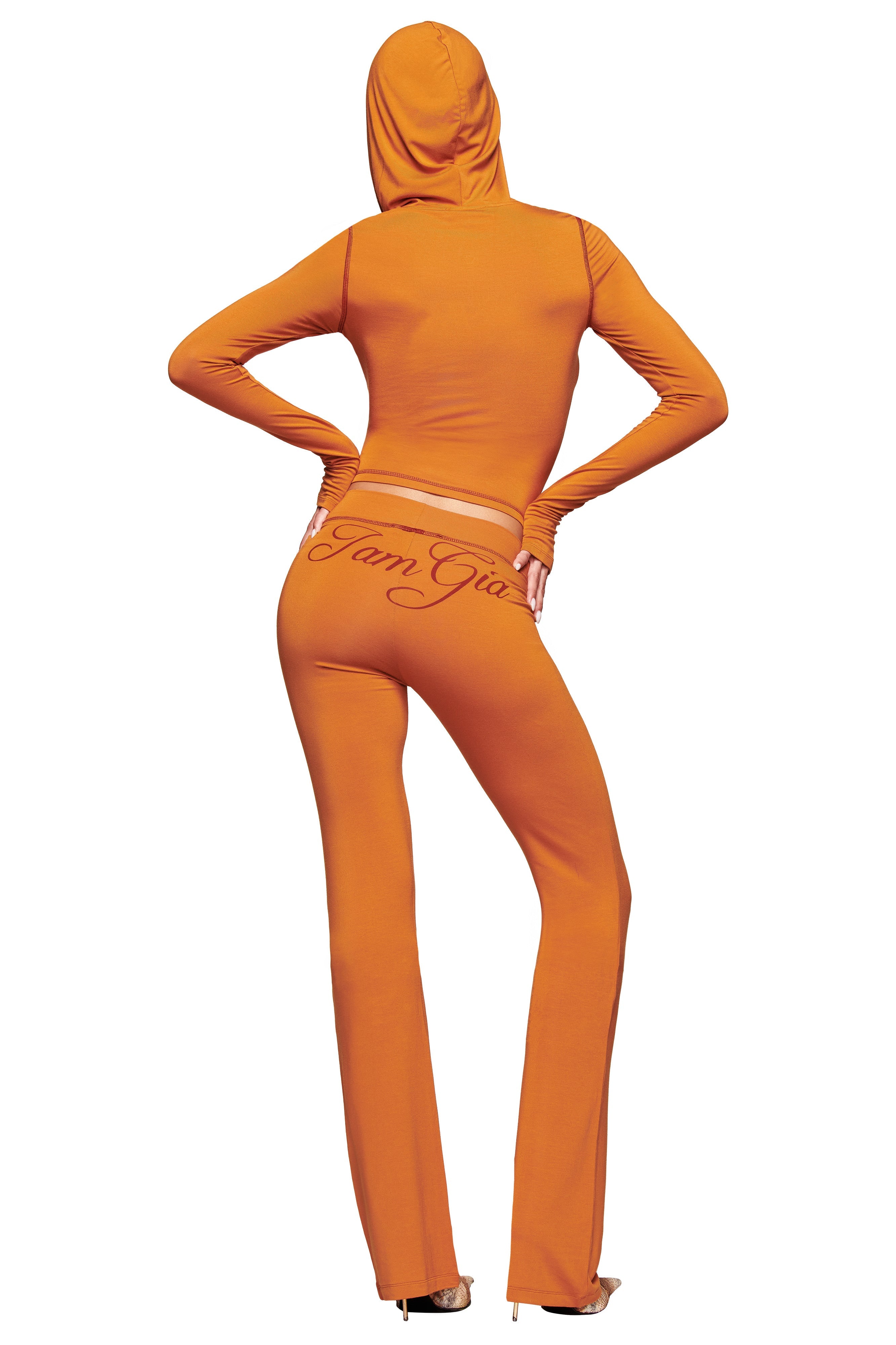 A person is seen from the back wearing I.AM.GIA's BLARE TRACKPANT - PUMPKIN with a matching pumpkin spice hooded crop top; flared pants feature cursive waistband text, and their heels are visible.