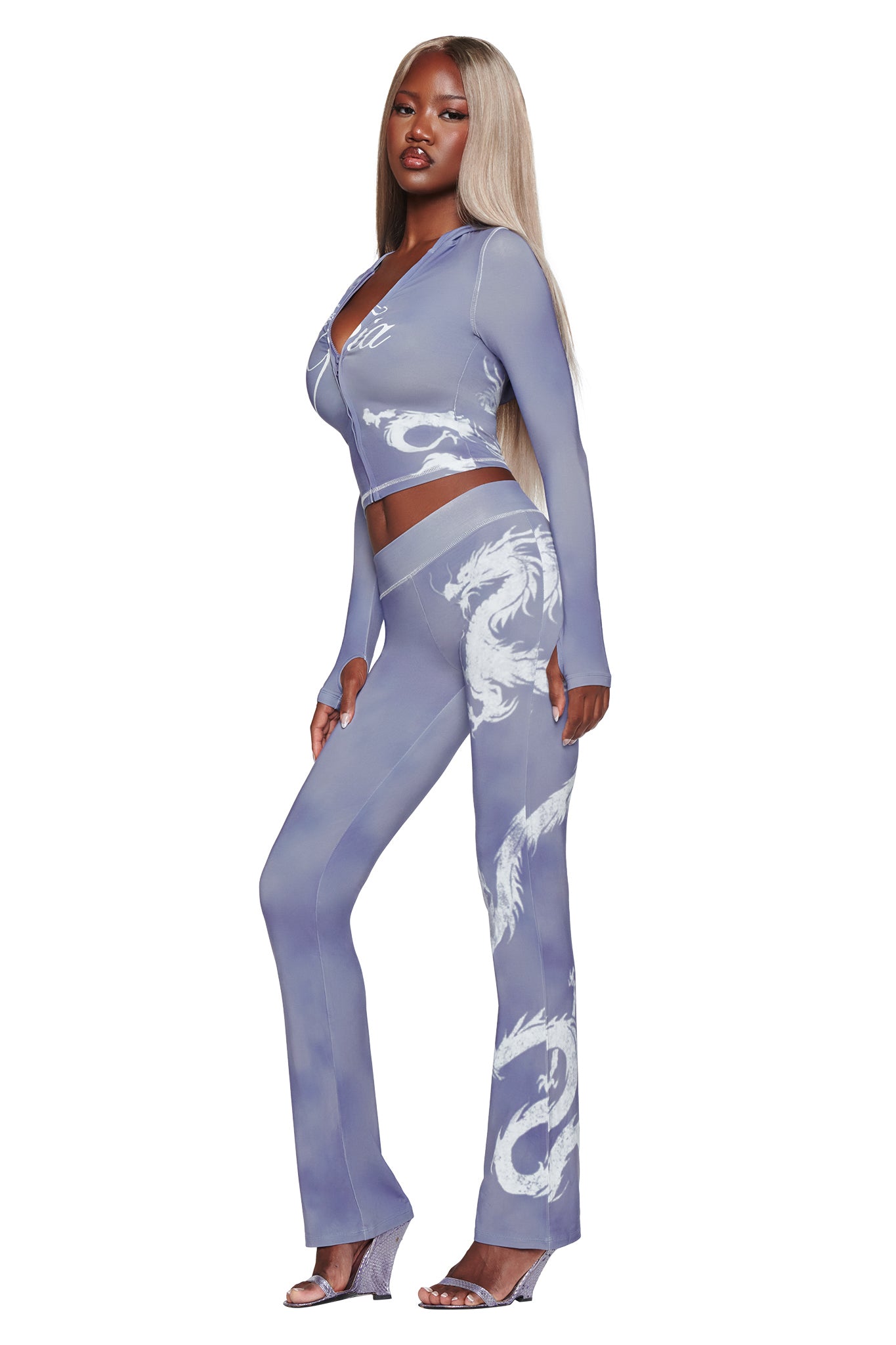 A woman with long blonde hair wears a fitted, light purple two-piece outfit with a white dragon design, paired with I.AM.GIA BLARE TRACKPANT in steel blue dragon print. She stands sideways in clear high-heeled sandals against a white background.