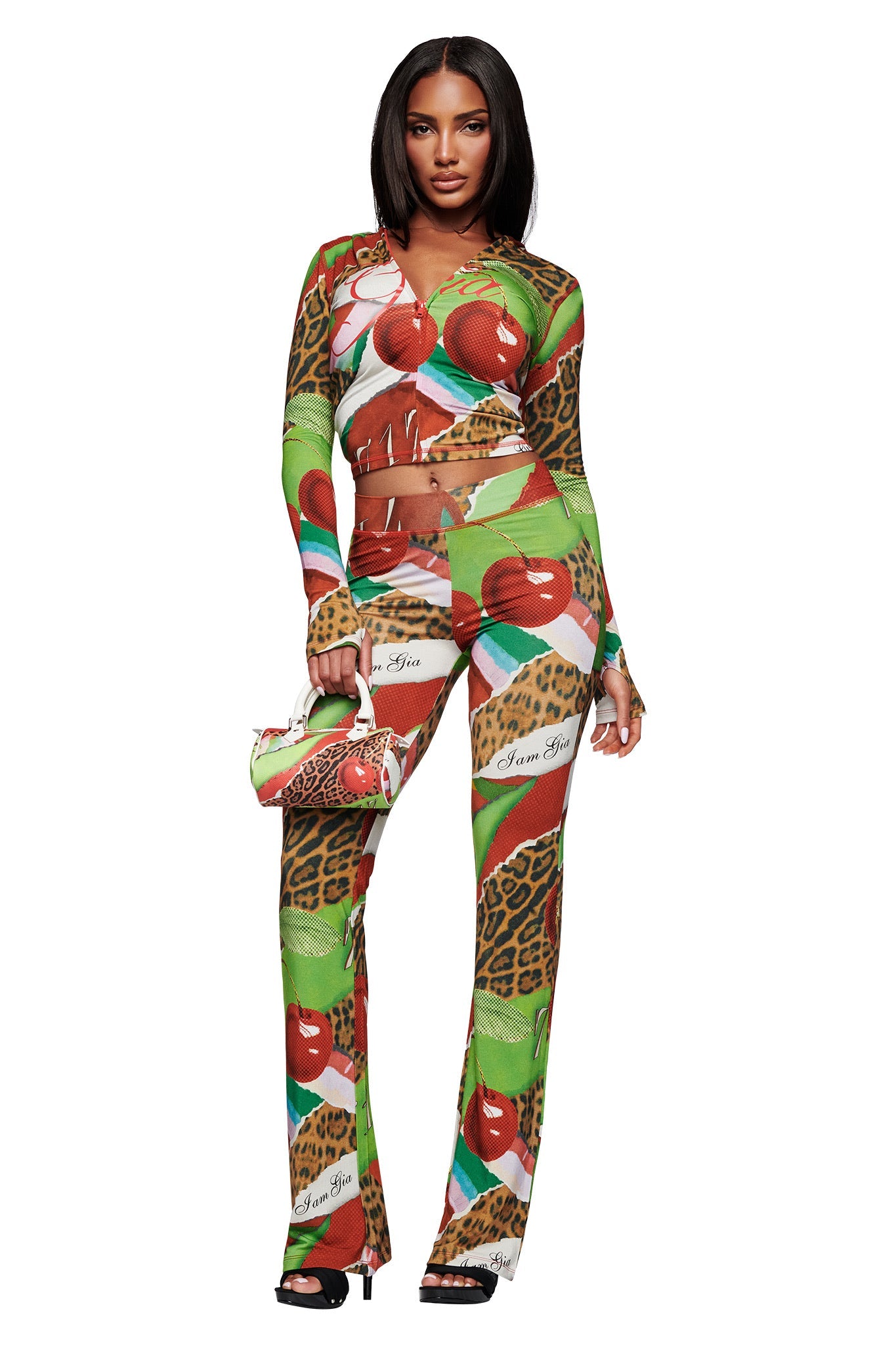 A woman with straight shoulder-length hair wears I.AM.GIA’s BLARE HOODIE in the vivid Wild Cherry print, featuring apples, leopard spots, and hearts, paired with black heels and a matching handbag, standing against a white background.