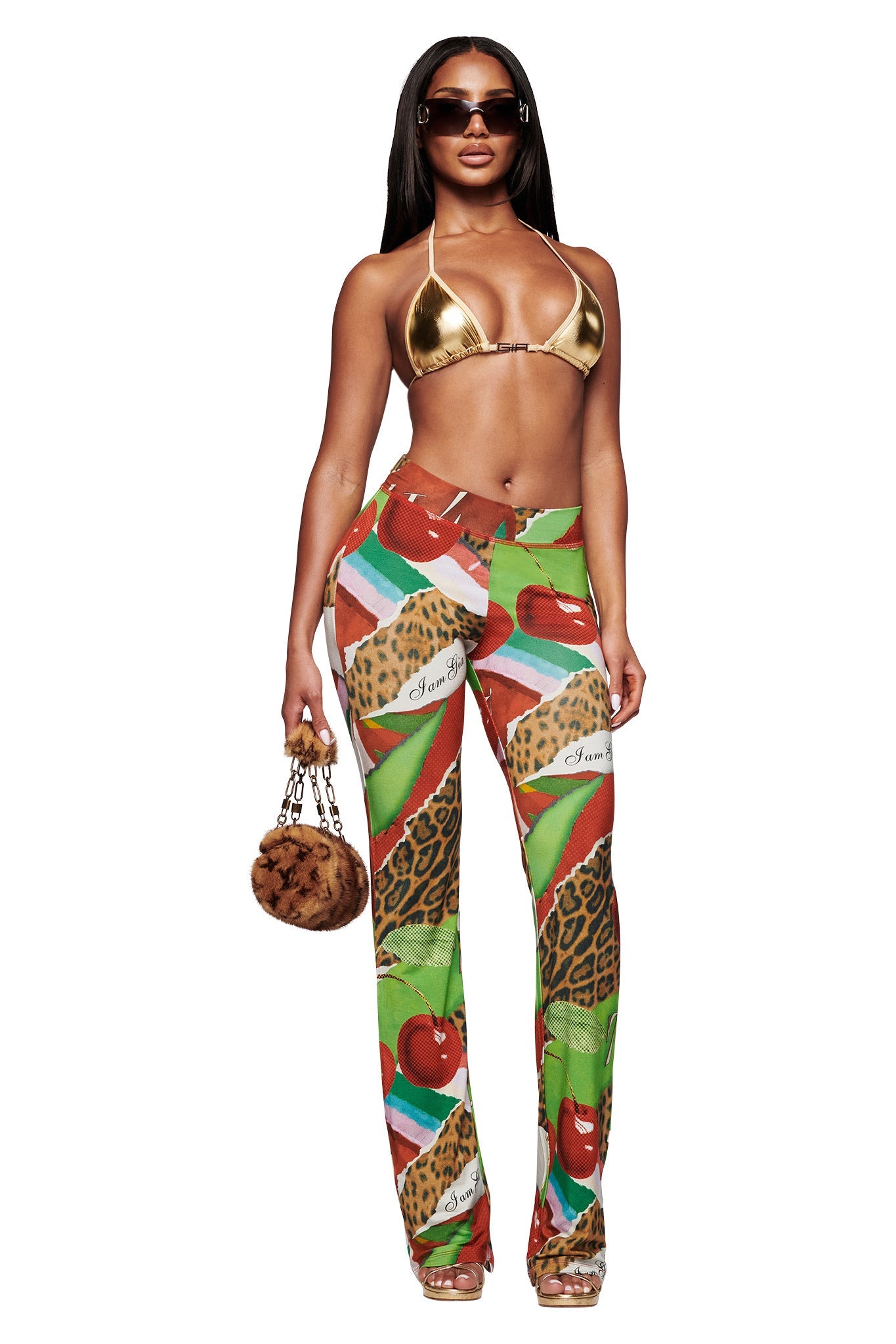 A woman poses in I.AM.GIA's BLARE TRACKPANT - WILD CHERRY, featuring animal and fruit prints, styled with a gold bikini top, clear heels, sunglasses, and a small furry handbag against a white background.