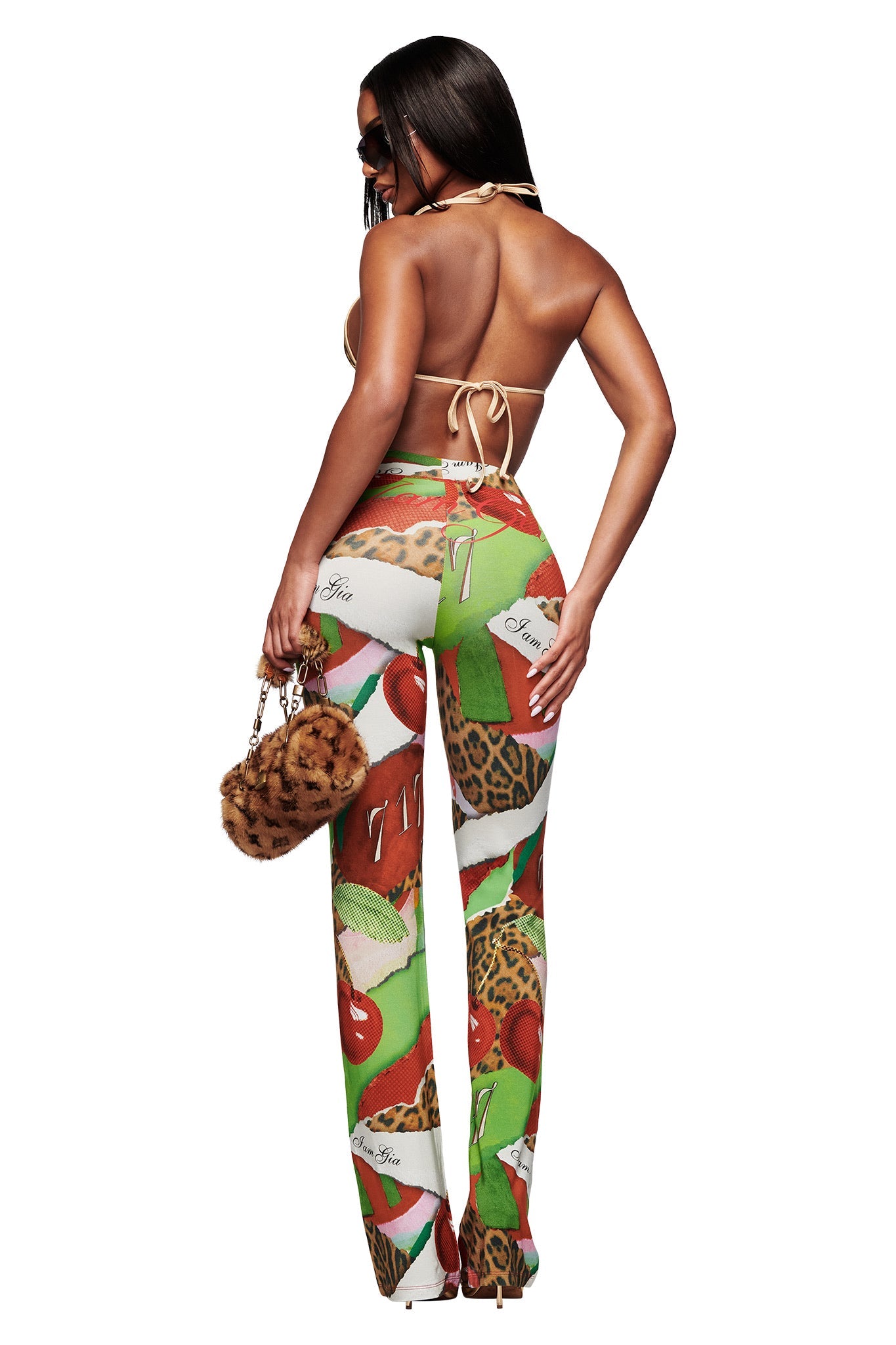 A woman faces away in the I.AM.GIA BLARE TRACKPANT - WILD CHERRY and a backless halter top, carrying an animal print purse and wearing sunglasses with heels. The background is plain white.