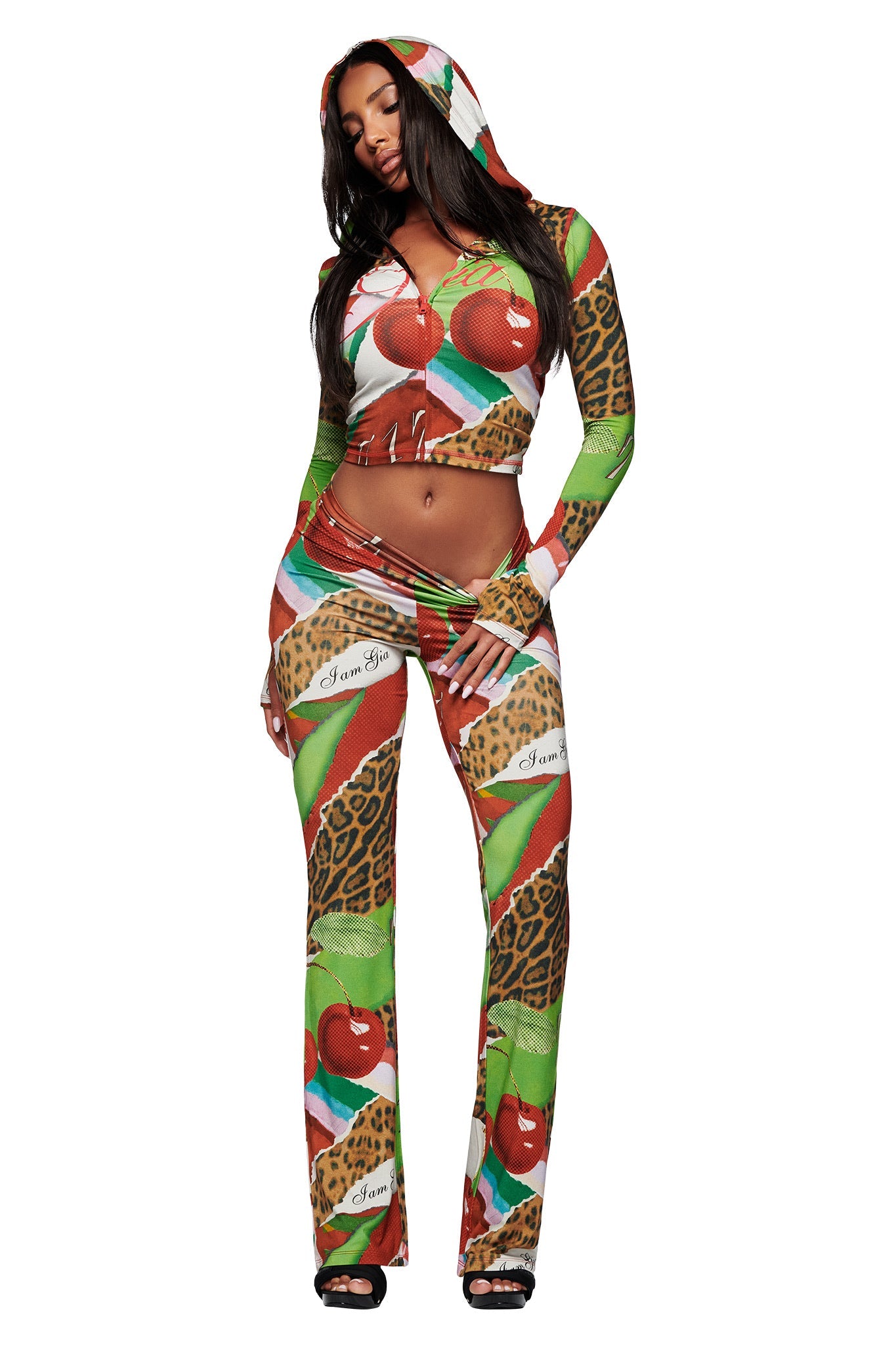 A woman models I.AM.GIA's BLARE TRACKPANT - WILD CHERRY set, a vibrant, form-fitting bamboo outfit with a hood, bold animal prints, red apples, green leaves, script text, a crop top, and flared track pants.