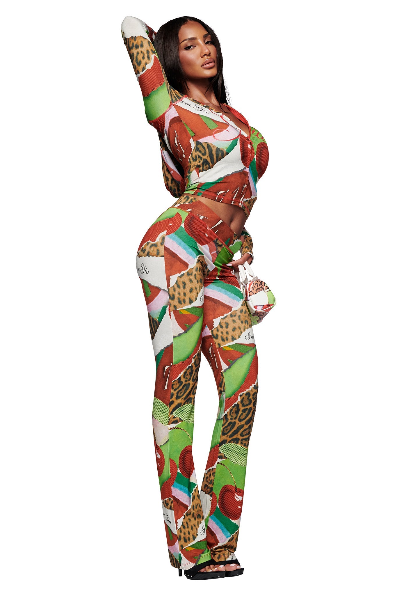 A woman poses in a vibrant I.AM.GIA outfit, featuring the BLARE TRACKPANT - WILD CHERRY and a matching long-sleeve, slightly cropped top with wild cherry print, animal motifs, and geometric patterns. She completes the look with open-toe sandals.