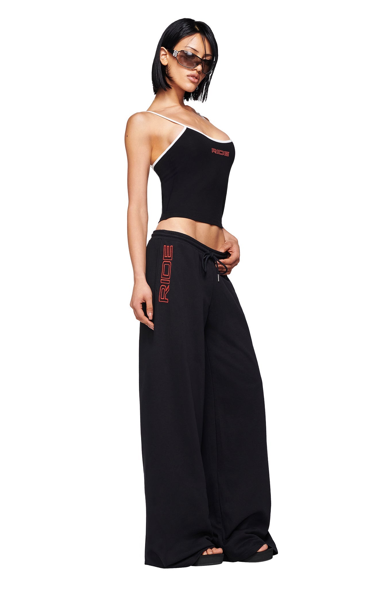A woman poses confidently in sunglasses, a black asymmetrical "RIDE" tank, and I.AM.GIA's JUSTINE PANT - BLACK: exclusive baggy track pants with a low-rise waist and vertical red "RIDE" lettering, set against a white background.