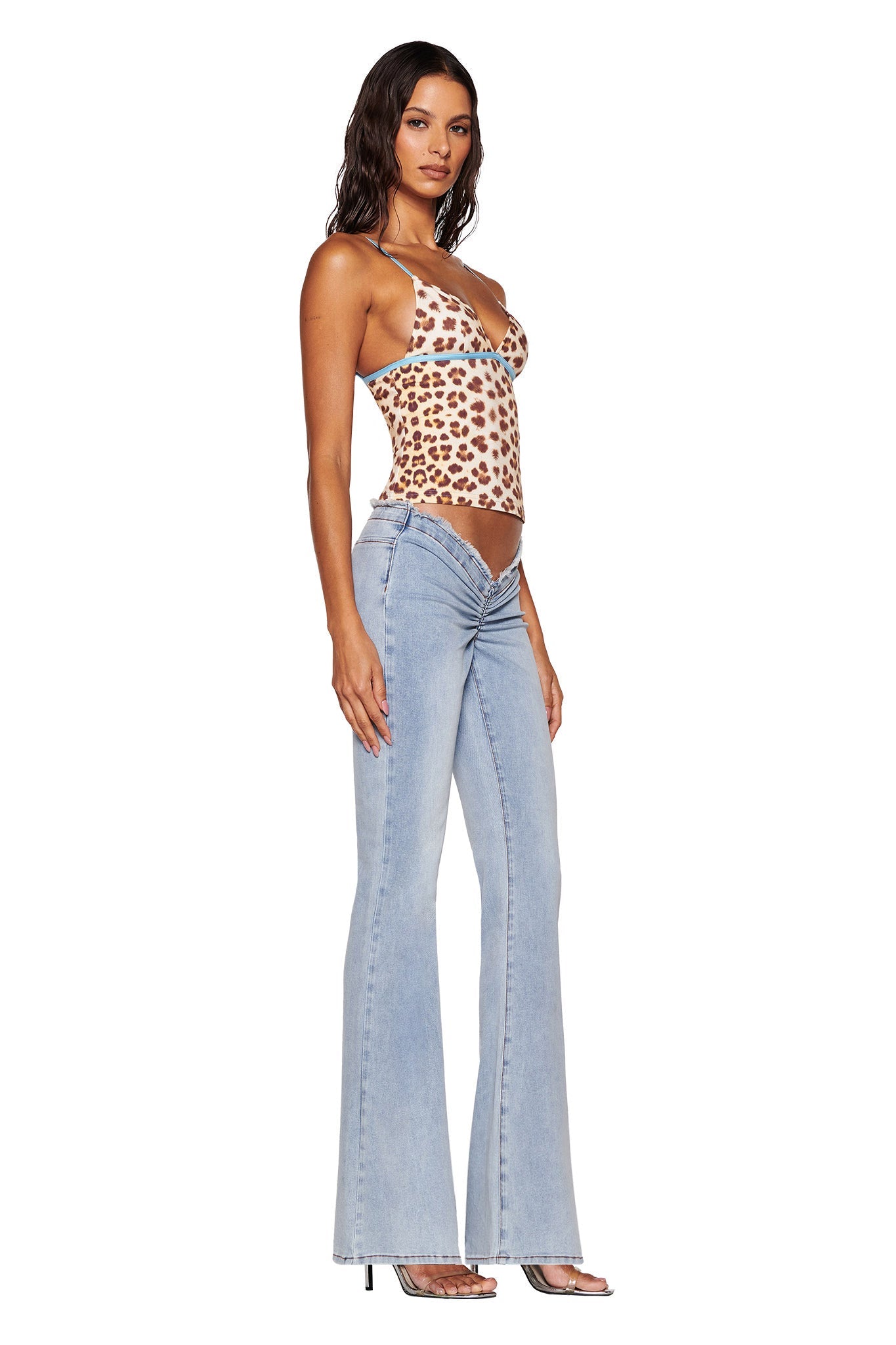 A person wearing the EDITA TOP in Neutral Leopard Wild Card by I.AM.GIA and light blue flared jeans stands against a white background, highlighting their high-heeled sandals.