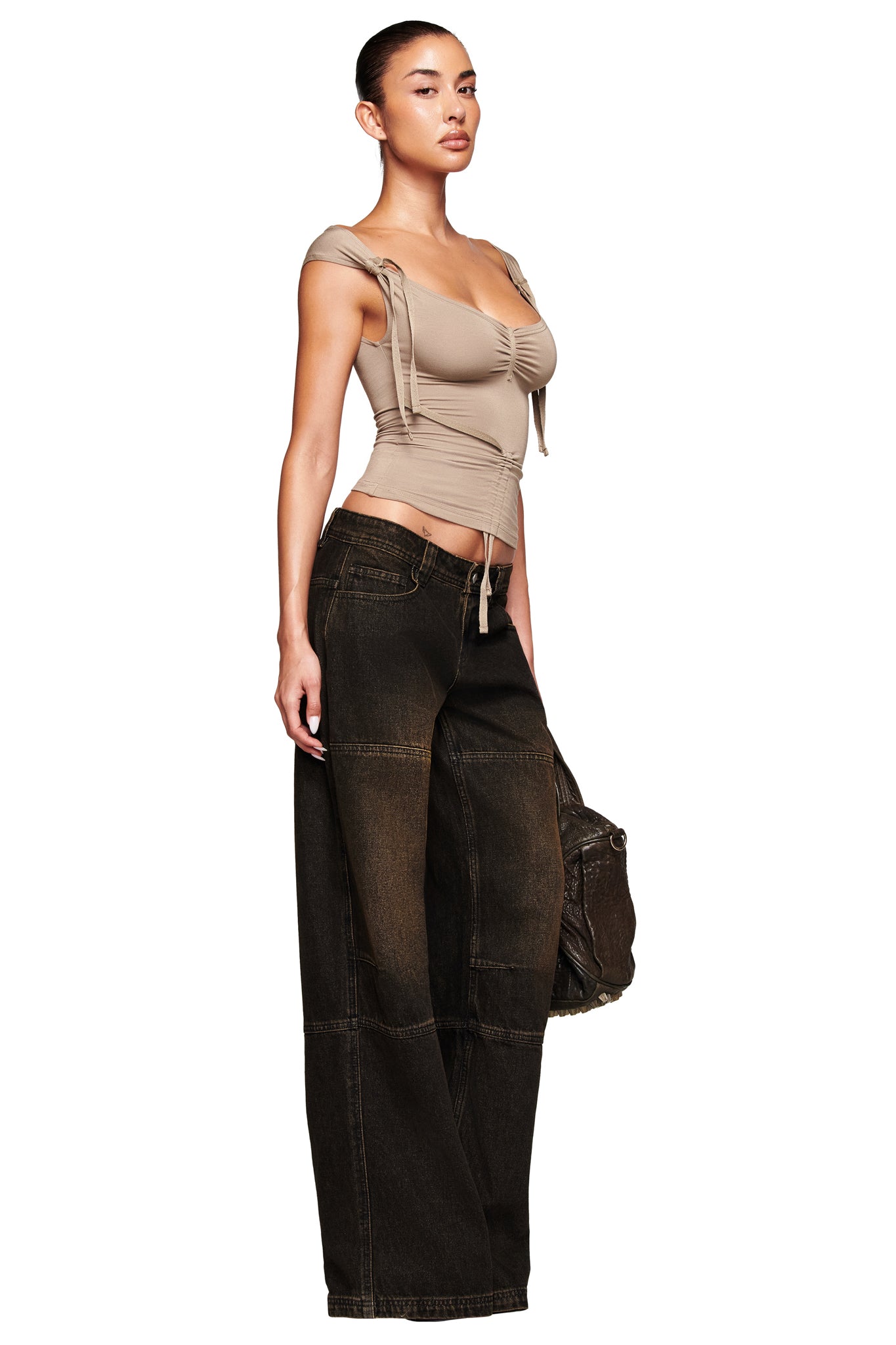 A woman stands confidently in I.AM.GIA's MISTY JEAN - BLACK, paired with a fitted beige ruched top and holding a large textured bag by her side.