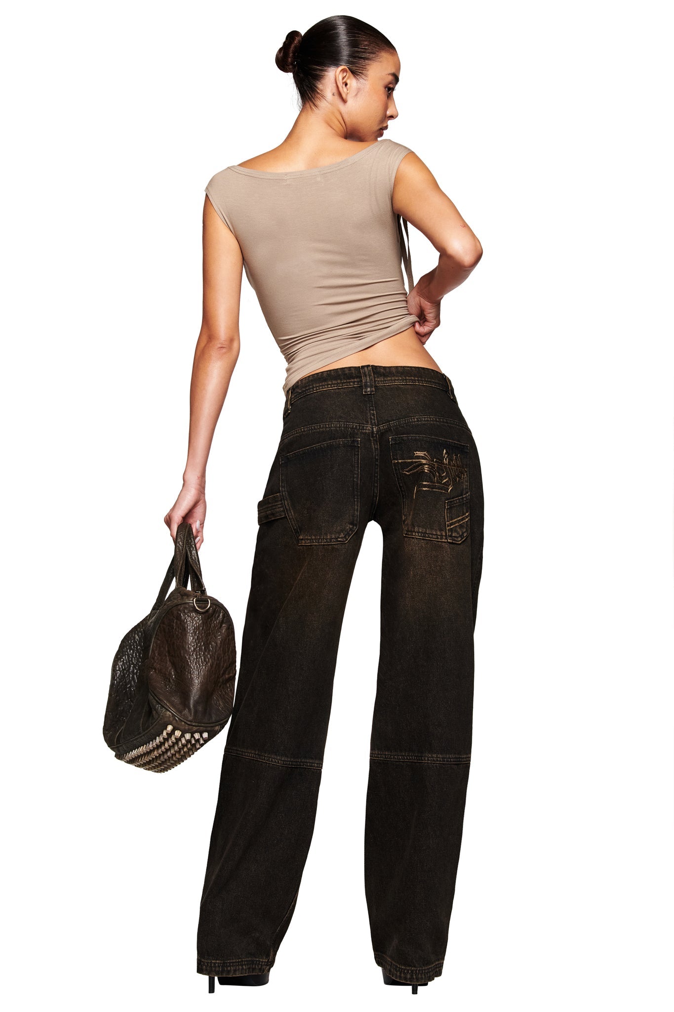 A woman with dark hair in a bun wears a fitted beige top and the I.AM.GIA MISTY JEAN - BLACK in a baggy fit, holding a studded brown textured handbag while posing against a white background and glancing over her shoulder.