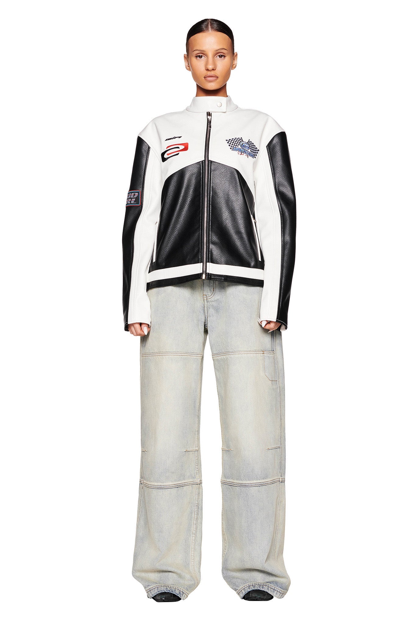 A person with a slicked-back hairstyle wears an oversized black and white I.AM.GIA jacket adorned with various patches, paired with wide-legged MISTY JEAN - BLUE : VINTAGE WASH jeans from I.AM.GIA. They stand facing forward against a plain white background.