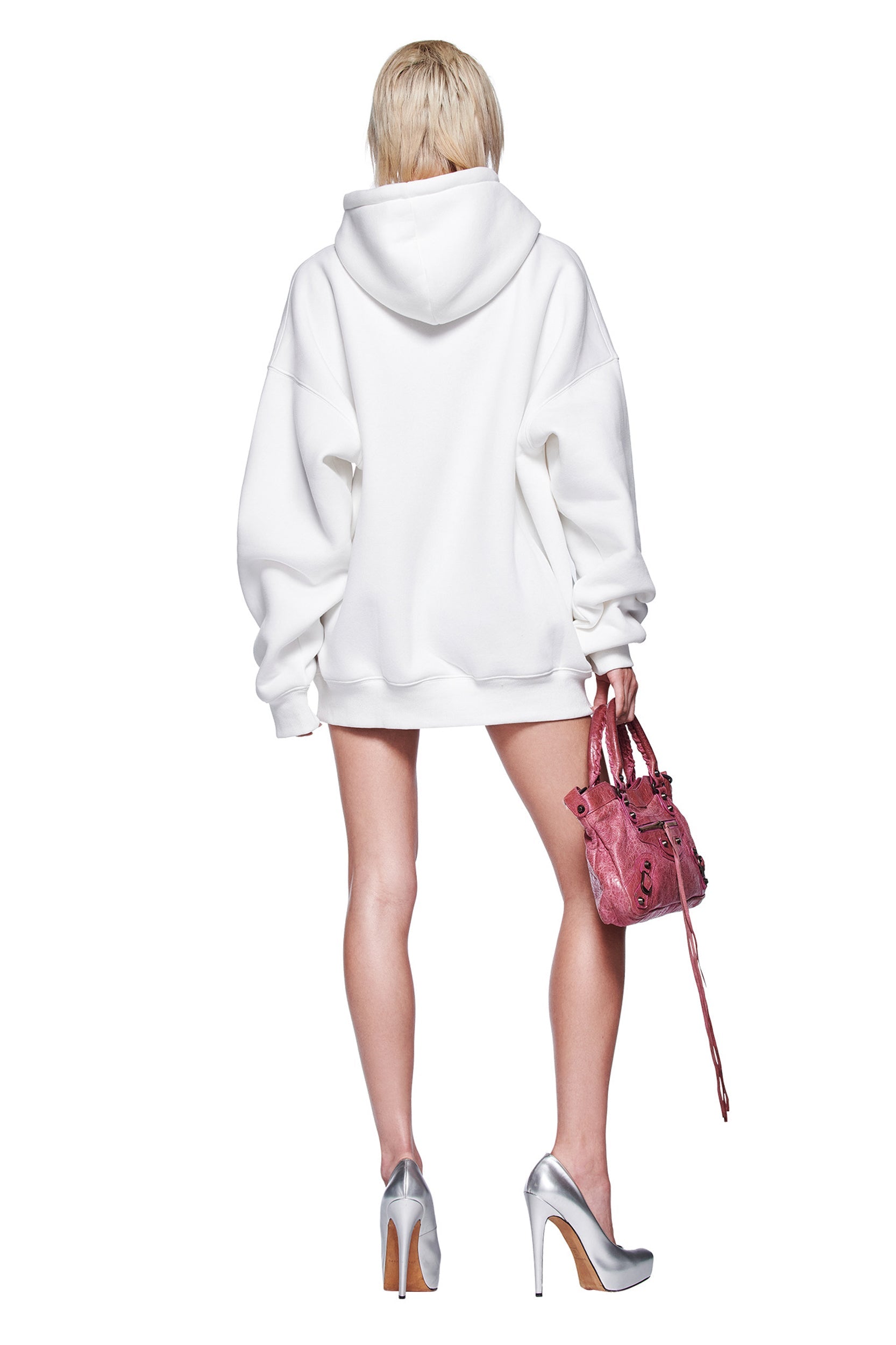 A person with short blonde hair faces away, modeling the oversized I.AM.GIA DORIAN HOODIE - WHITE, paired with a small pink handbag and silver high heels. The plain white background creates an airbrushed artwork effect.