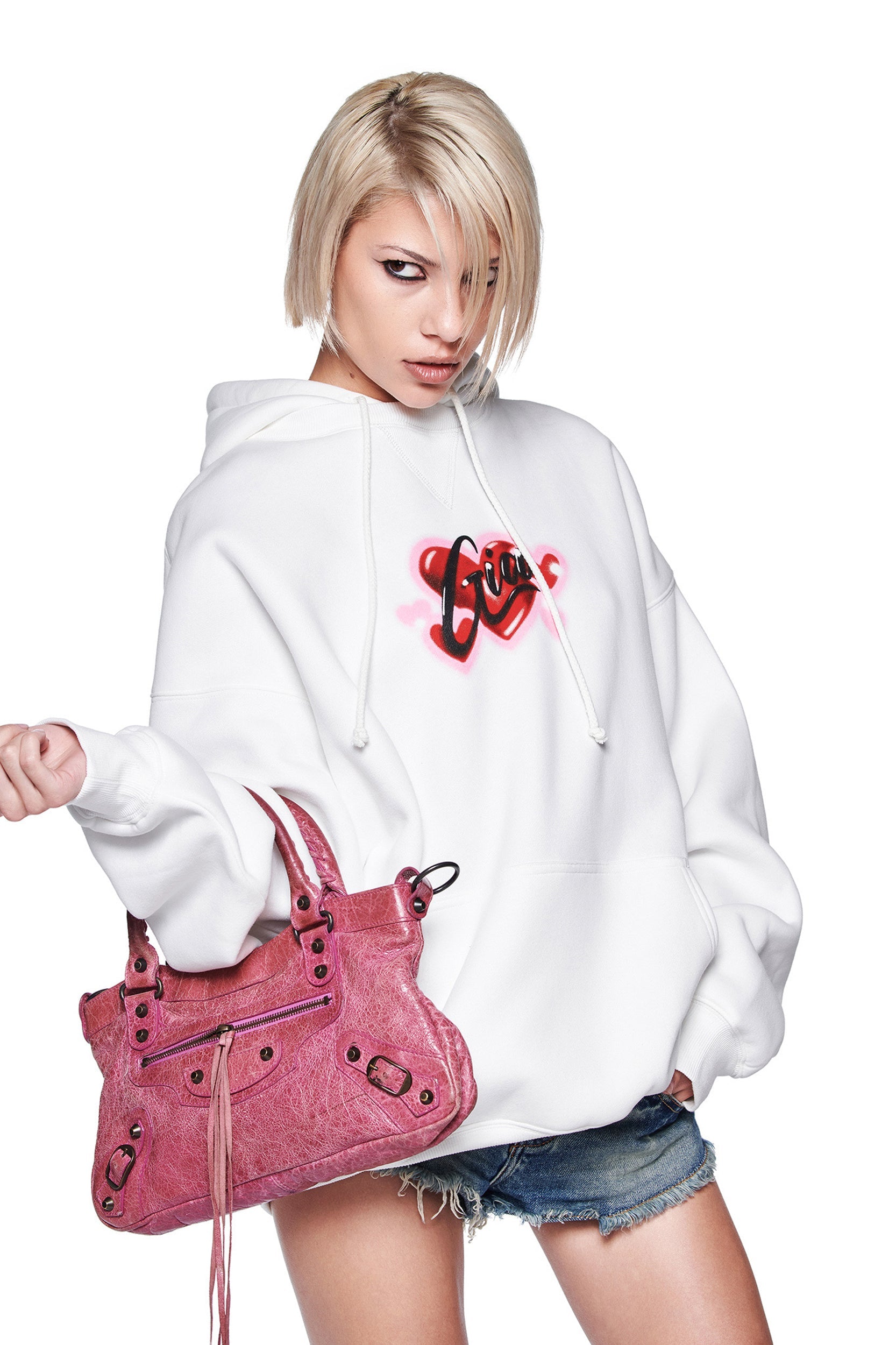A person with short blonde hair wears the oversized I.AM.GIA DORIAN HOODIE in white, featuring a red heart and "Love" airbrush design, styled with blue denim shorts and a pink handbag against a white background.