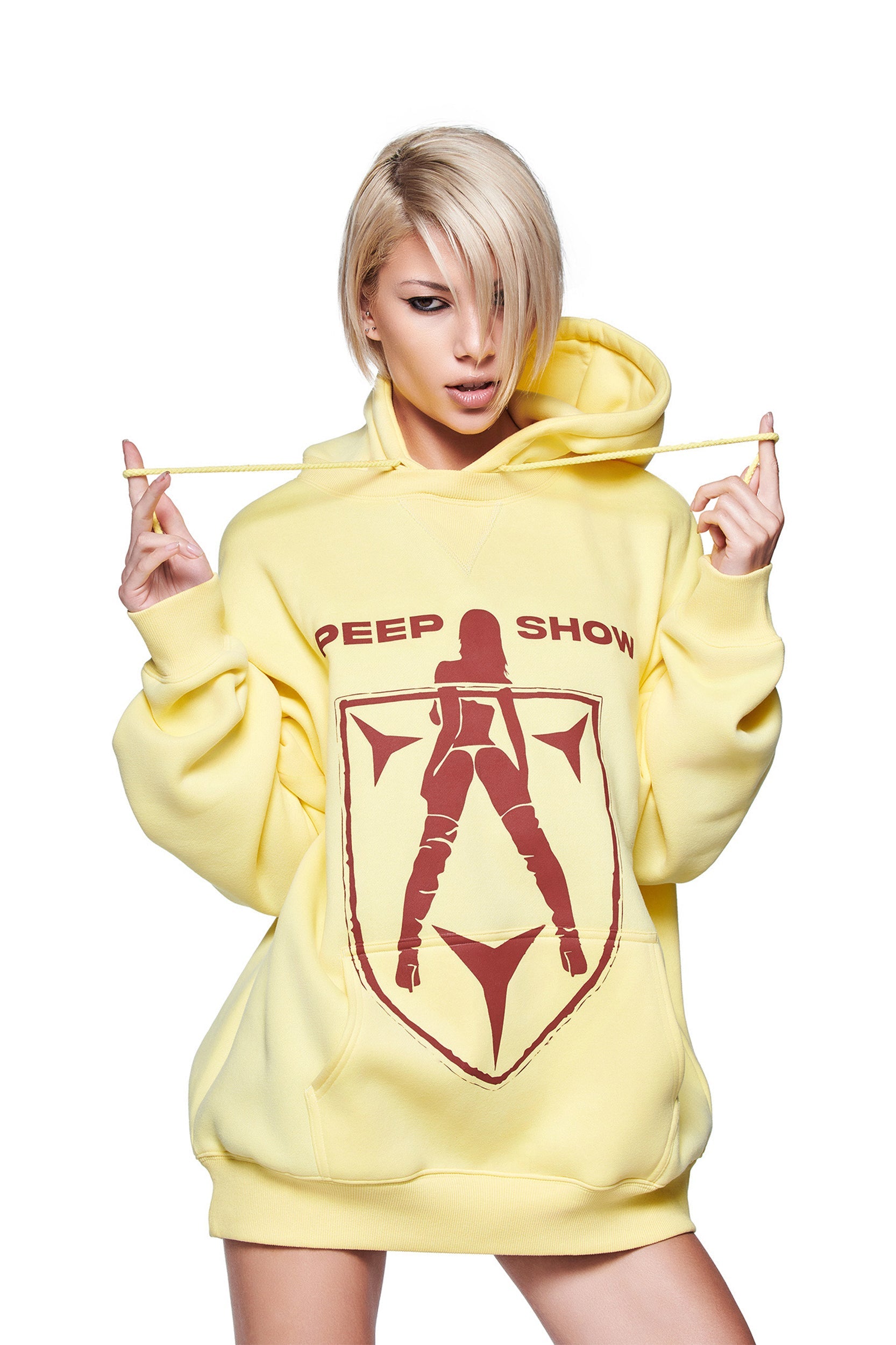 A person with short blonde hair models the oversized I.AM.GIA DORIAN HOODIE in yellow, featuring "PEEP SHOW" and custom GIA artwork, posing while holding the hoodie strings against a white backdrop.