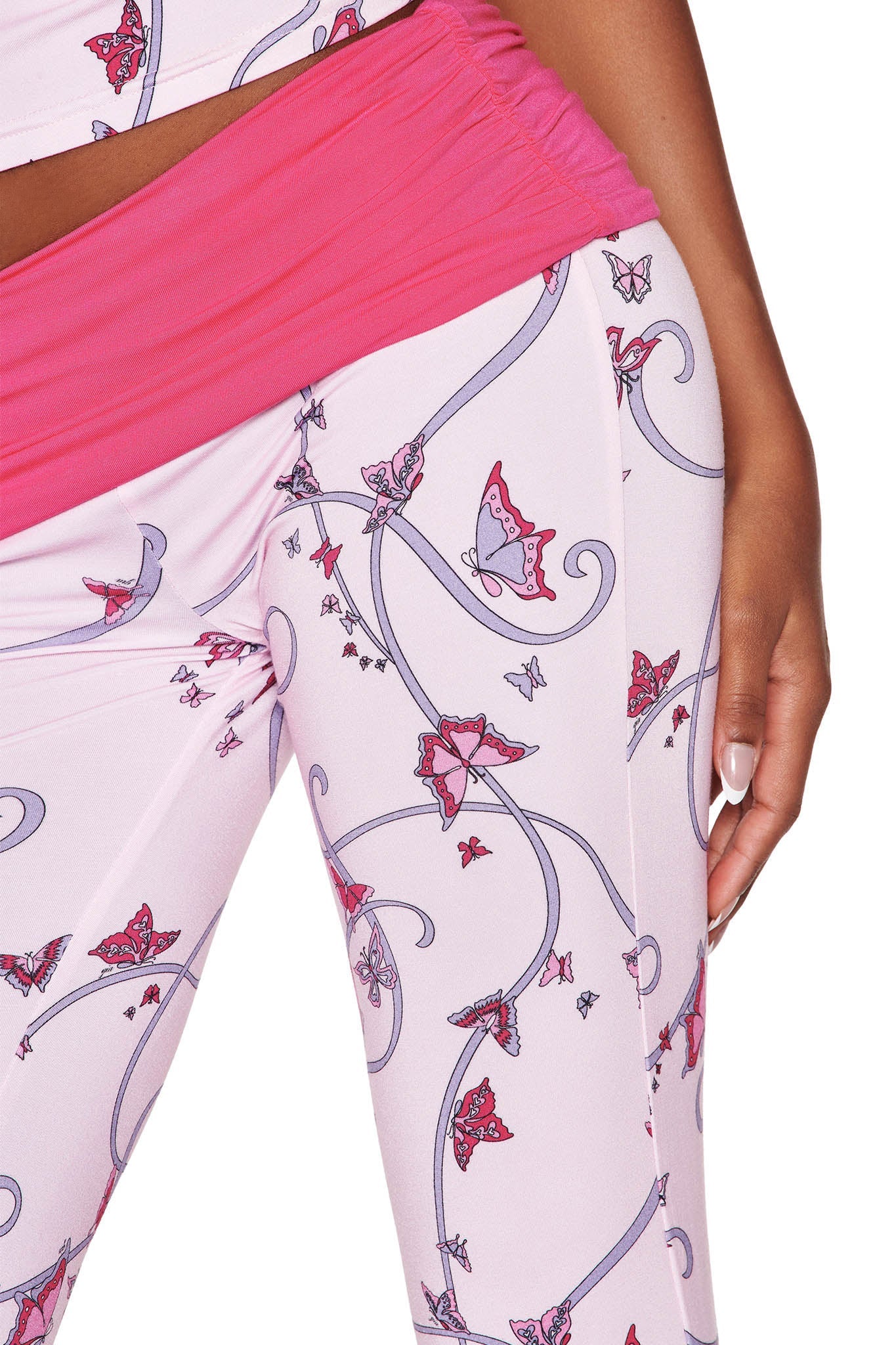 The person is wearing I.AM.GIA's JANNIS PANT in light pink, featuring a mid-rise waist and an enchanting butterfly ribbon print with swirly lines, paired with a bright pink top partially visible, while their hand rests gracefully by their side.