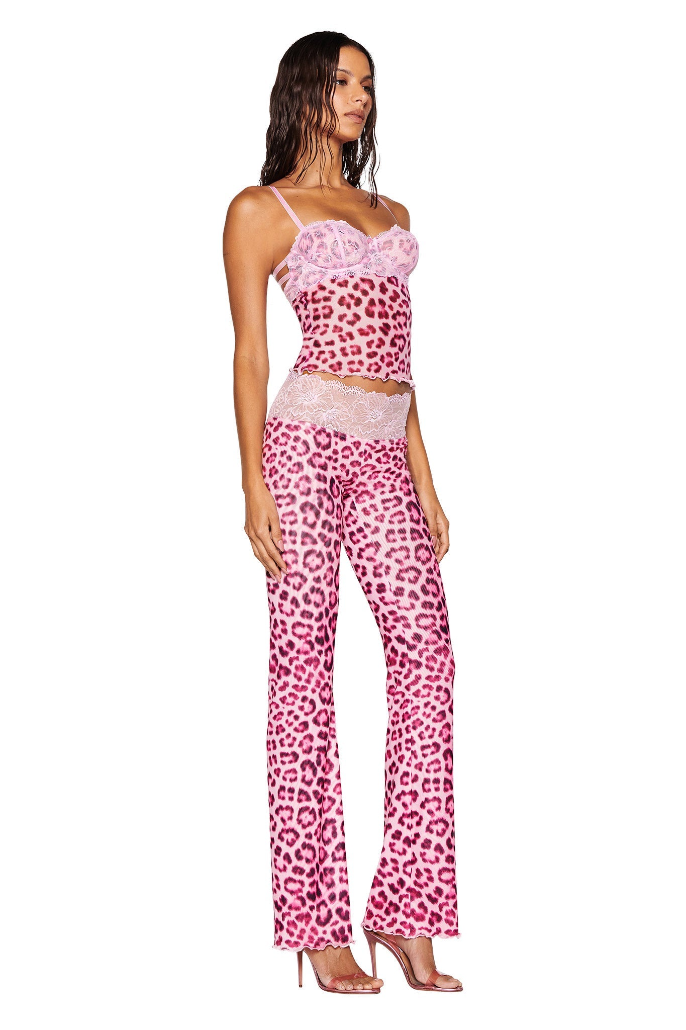 A person confidently poses in the I.AM.GIA SILO TOP - PINK LEOPARD outfit, featuring lace details with a camisole and matching flared pants. Styled with wet-look hair and high-heeled shoes, the ensemble includes a hint of mesh fabrication for added allure.