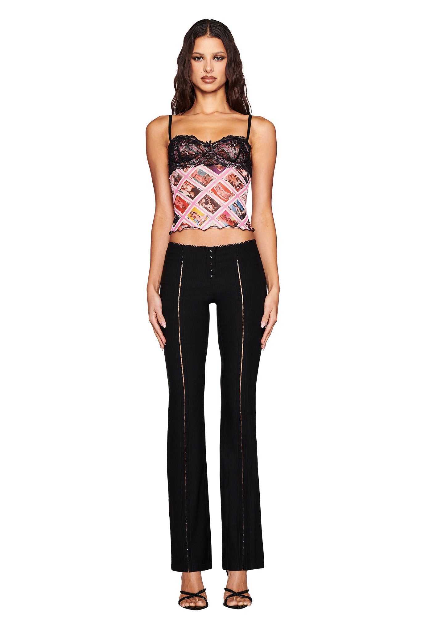 A woman with long, wavy dark hair stands facing forward in I.AM.GIA's SILO TOP - MULTI : GIA FACE CARD, paired with black pants featuring visible stitching and black high-heeled sandals. The background is plain white.