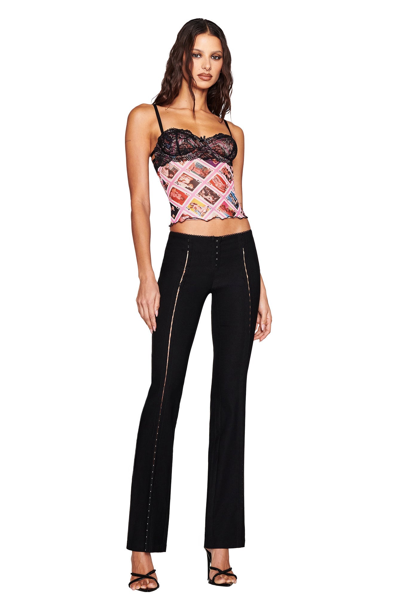 A woman confidently stands with hands to her sides, wearing the I.AM.GIA SILO TOP in a colorful design with lace trim, paired with black fitted pants and black high-heeled sandals. Her long, wavy hair cascades over the plain white background.