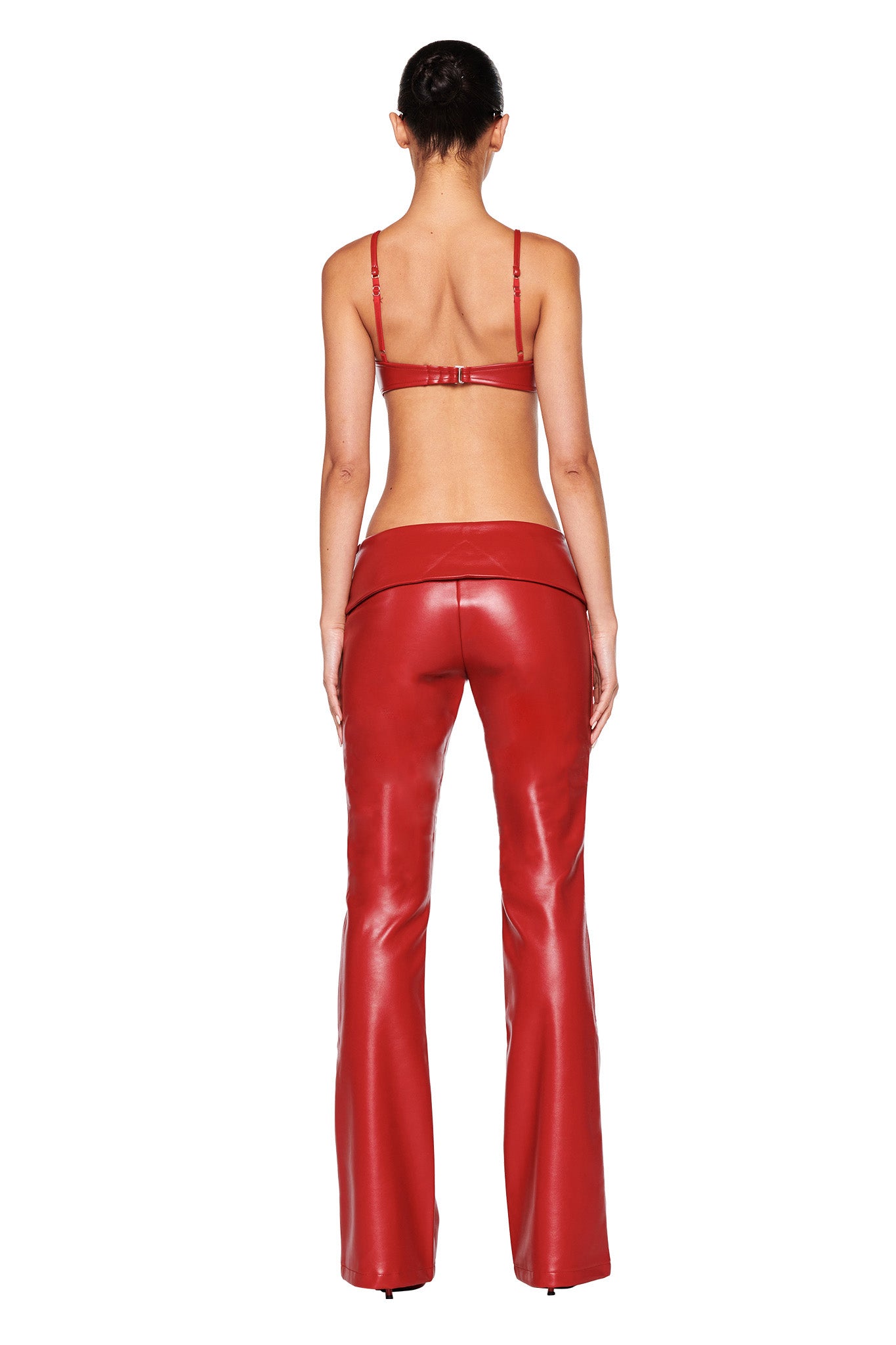 A person stands facing away, wearing a red I.AM.GIA bra and the AMALTHEA PANT 2.0 in red. Their hair is tied back, and the glossy pants add a striking finish. The photo captures the full length of the person from head to toe against a plain white background.