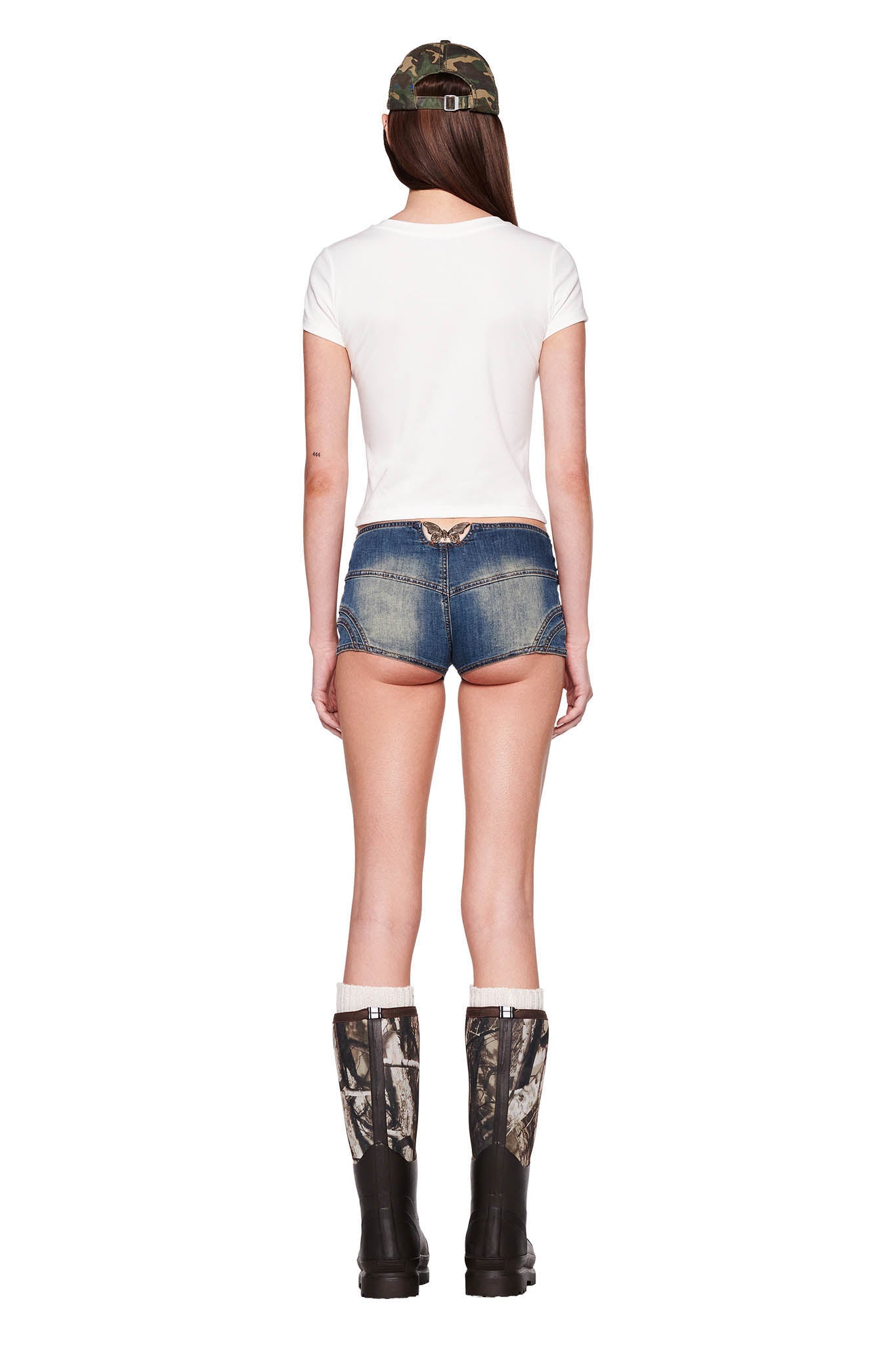 A woman stands with her back to the camera wearing the ODEZIA TOP - WHITE from I.AM.GIA, blue denim shorts, and tall black boots with camo patterns from I.AM.GIA. She sports a matching camo baseball cap, partially tucking her long brown hair under it. The background is plain white.