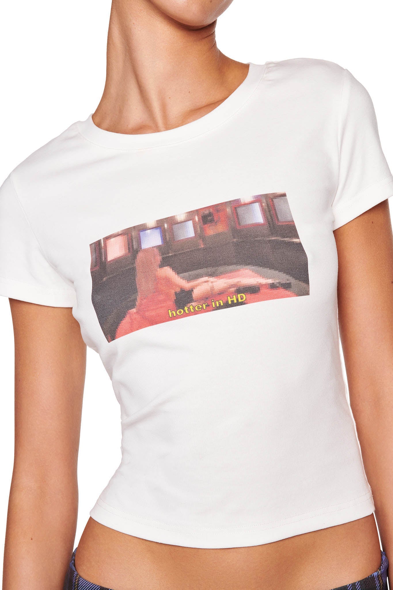 A person wearing the ODEZIA TOP - WHITE by I.AM.GIA, featuring a graphic of a scene with someone lying on a sofa surrounded by screens and the text "hotter in HD." The person wearing the shirt is visible only from the neck to the waist.