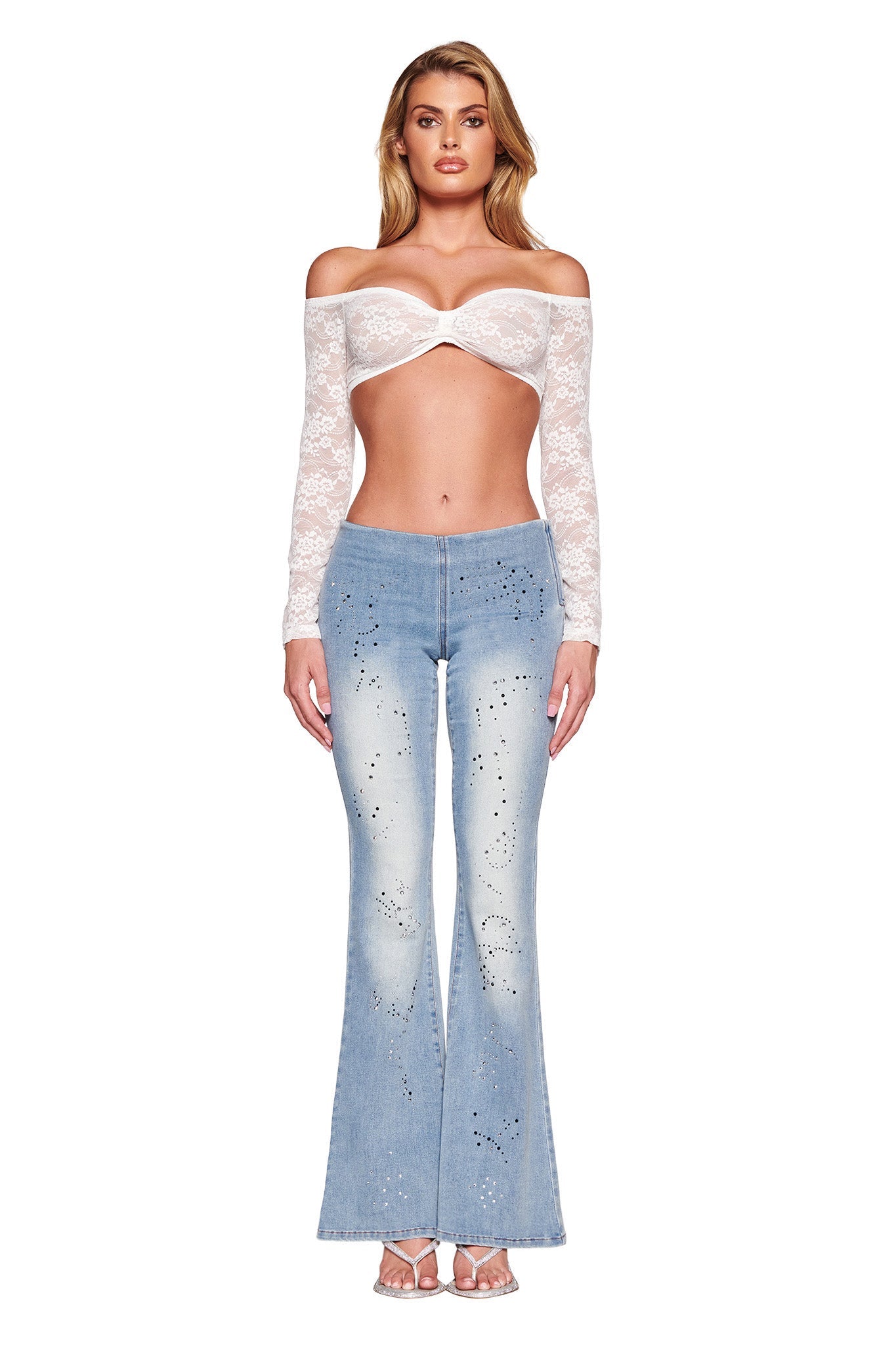 A person is standing and wearing a white, off-shoulder, long-sleeved lace top paired with the REQUIEM JEAN - BLUE from I.AM.GIA, featuring a high-waisted fit in light blue denim and a flared silhouette adorned with small diamante embellishments. They are also wearing open-toed high-heeled sandals. The background is white.