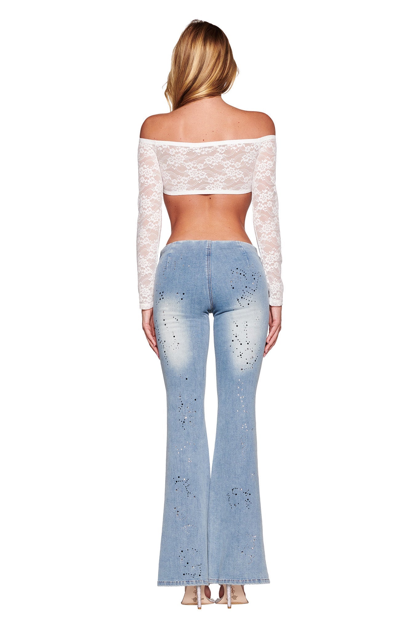 A person with long blonde hair is seen from the back, wearing an off-shoulder white lace crop top and light blue high-waisted REQUIEM JEAN - BLUE by I.AM.GIA. The jeans feature a flared silhouette with subtle diamante embellishments. They are standing against a white background, with their feet partially visible in beige heels.