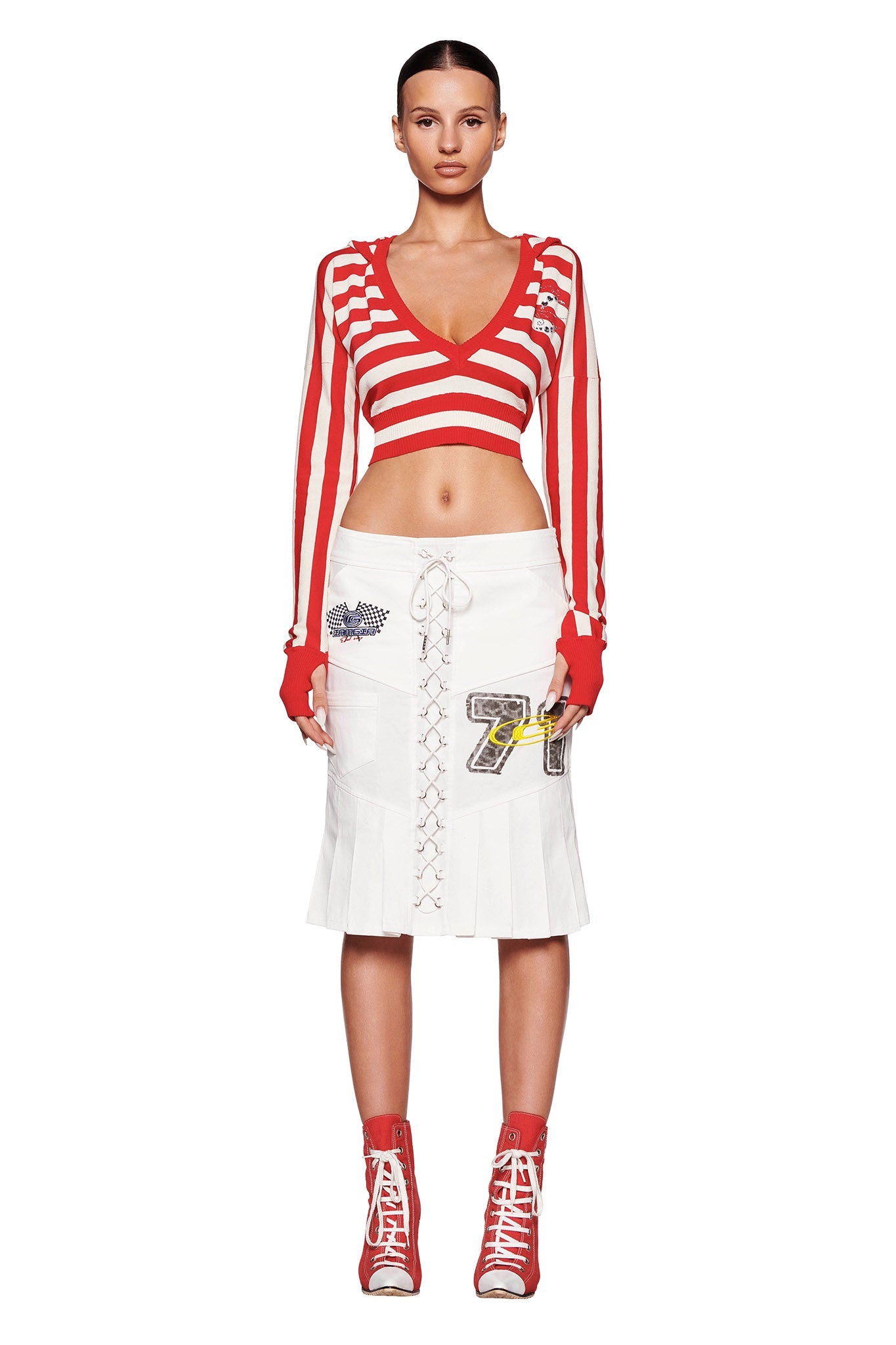 A model stands facing forward, showcasing the I.AM.GIA SUZI SWEATER in red stripe, paired with a white knee-length skirt featuring lace-up detailing and patches, and red high-top sneakers. The model's hair is styled in a sleek updo against a plain white background.