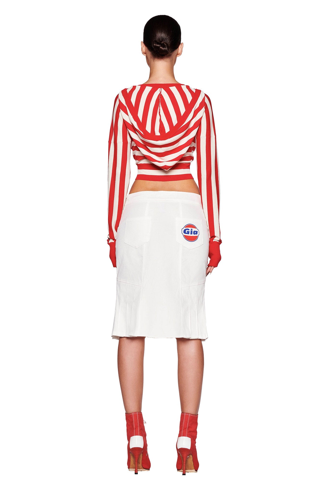 A person stands with their back to the camera, showcasing an I.AM.GIA SUZI SWEATER - RED : STRIPE long-sleeve crop top paired with a white skirt adorned with two logos on the back. They complete the look with red gloves, red high-heeled shoes, and hair styled in a sleek, tight bun. The background is plain white.
