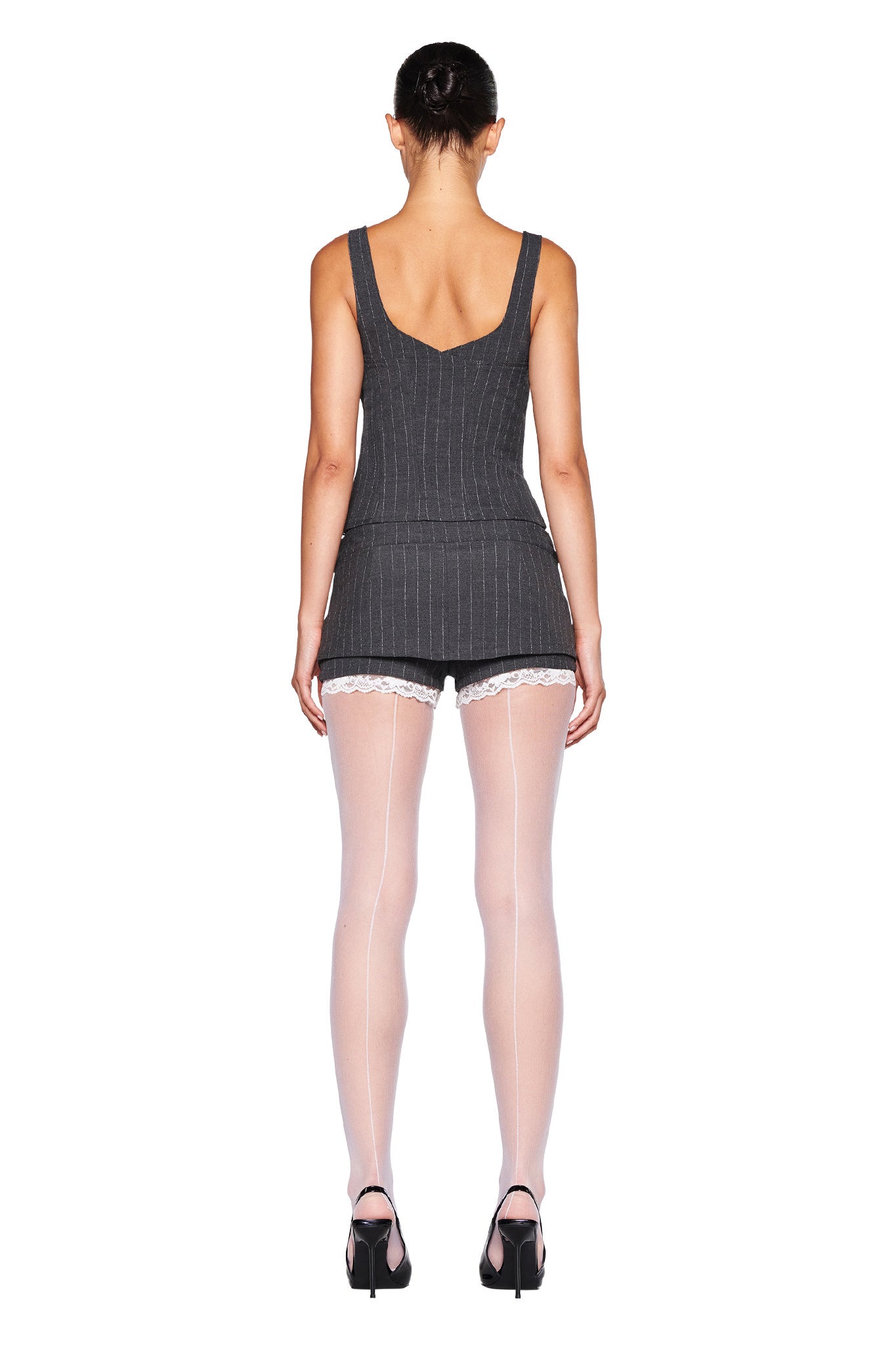 The image shows a person standing with their back to the camera, wearing a sleeveless, dark pinstripe top paired with the MARRION SKORT - GREY : PINSTRIPE by I.AM.GIA, which features built-in shorts. They are also wearing sheer white stockings with lace trim and black high-heeled shoes. Their hair is tied back in a bun.