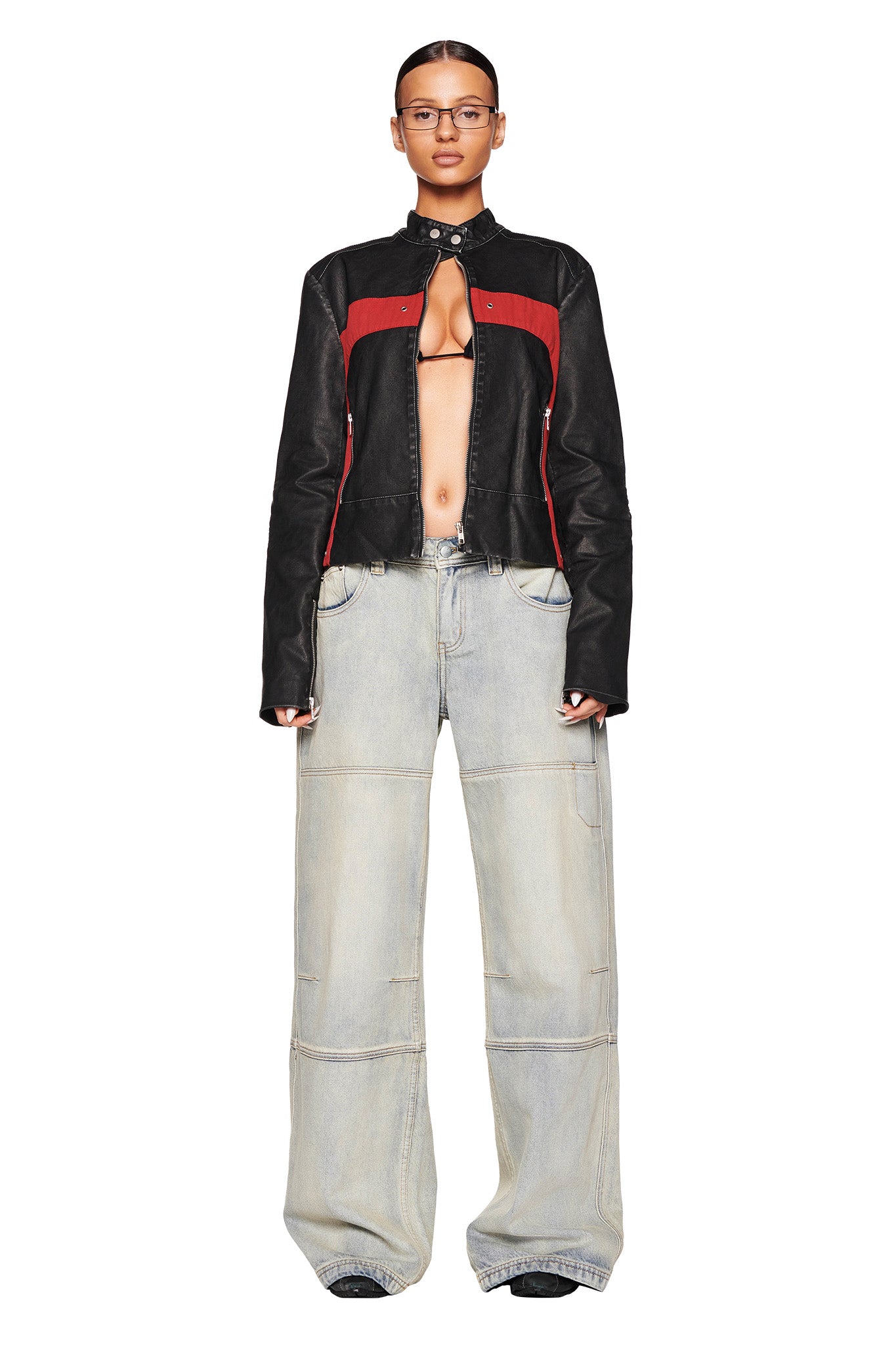 A person stands against a white background wearing a black and red leather I.AM.GIA jacket that is open in the front, revealing bare skin. They are also wearing the I.AM.GIA MISTY JEAN - BLUE : VINTAGE WASH, which are baggy, light-washed, wide-leg jeans that cover their shoes. They have a serious expression on their face.