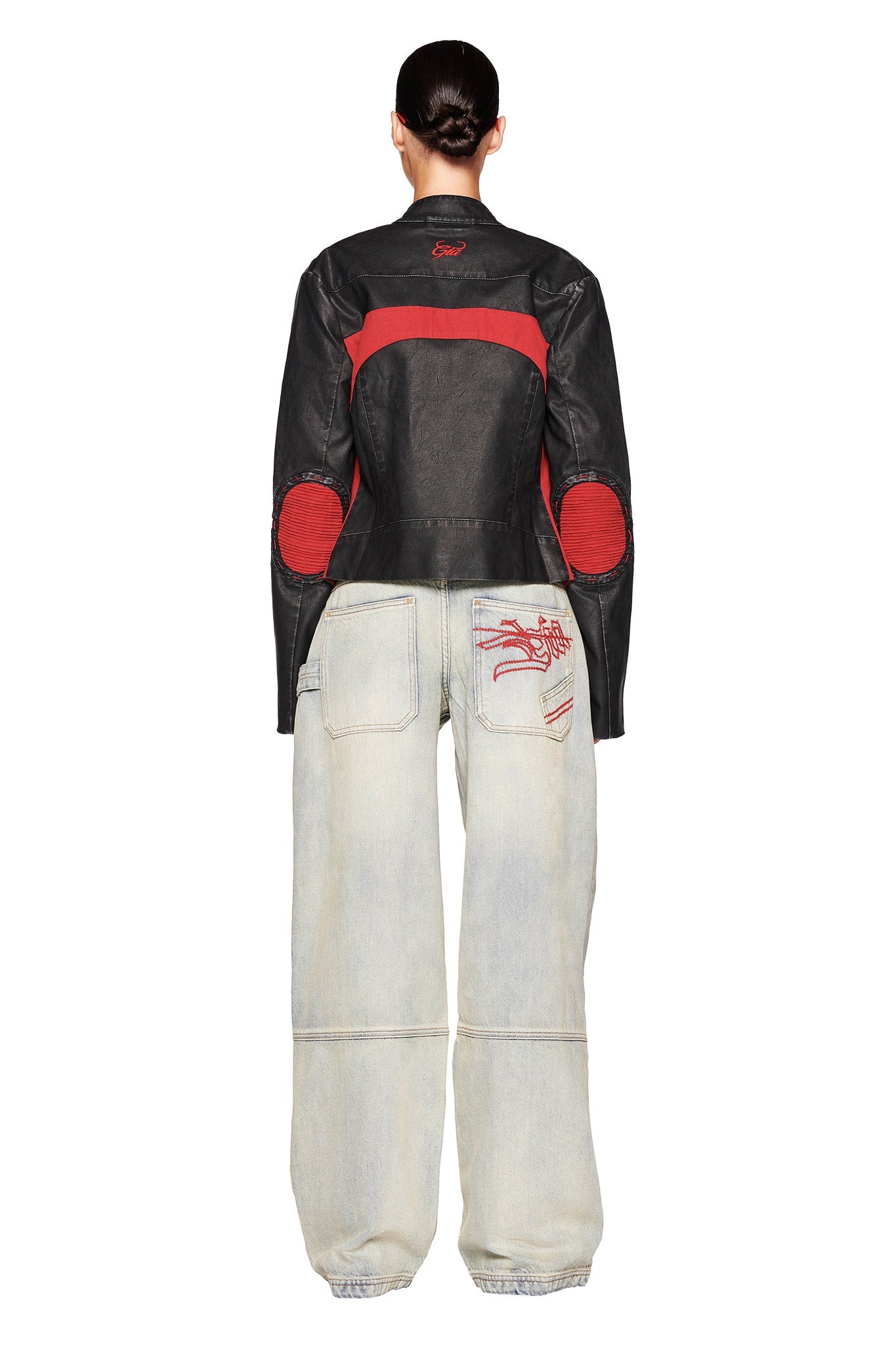 A person is standing with their back to the camera wearing a black and red leather jacket by I.AM.GIA, paired with the MISTY JEAN - BLUE : VINTAGE WASH from the same brand. Their hair is tied back, and the jacket has red patches on the elbows and shoulders. The jeans feature a red embroidered design on the back pocket.