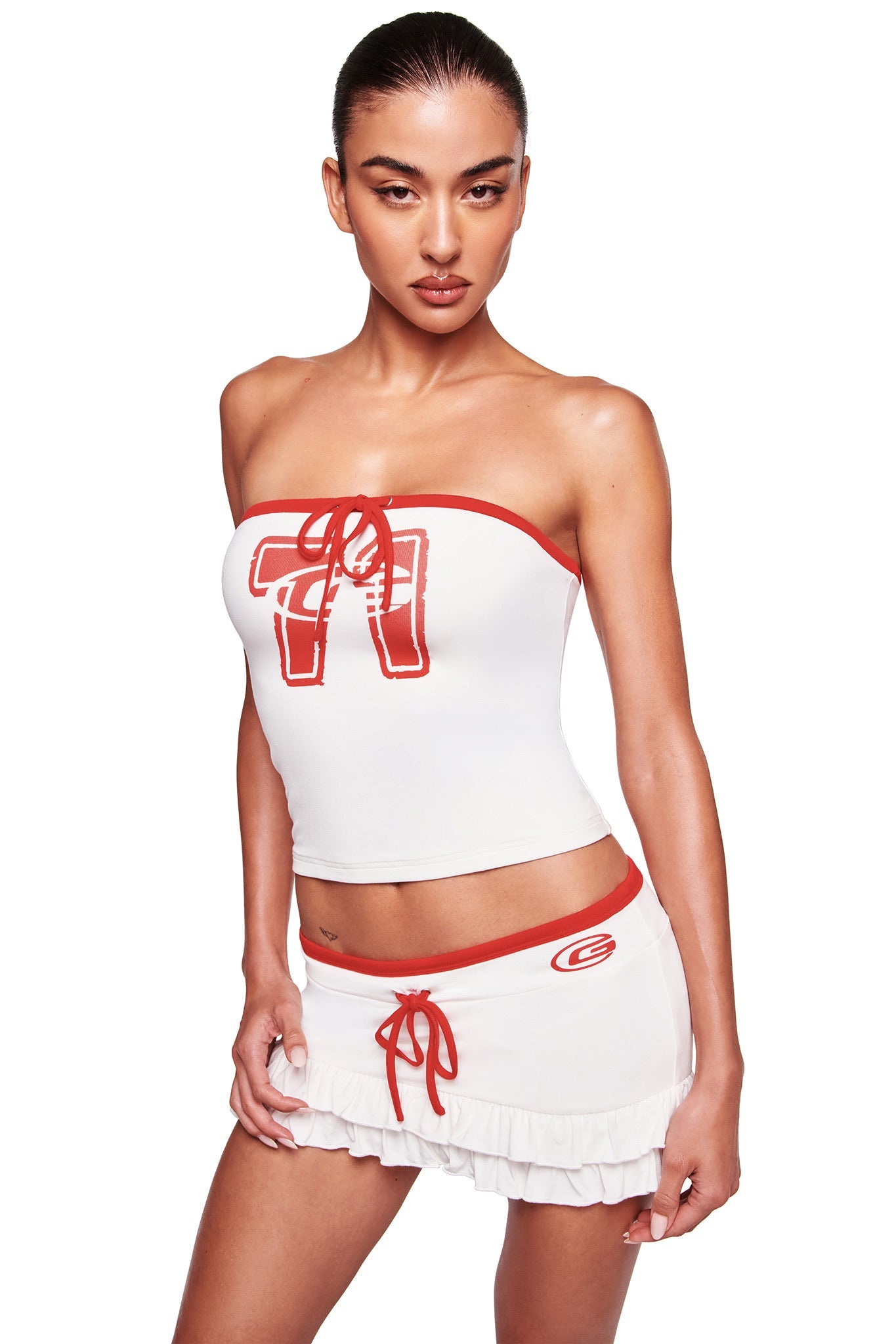 A woman models the I.AM.GIA MICHELLE BOOB TUBE in white, featuring bold red "71" graphics and trim, paired with a short ruffled skirt with a red waistband and emblem, set against a plain white background.