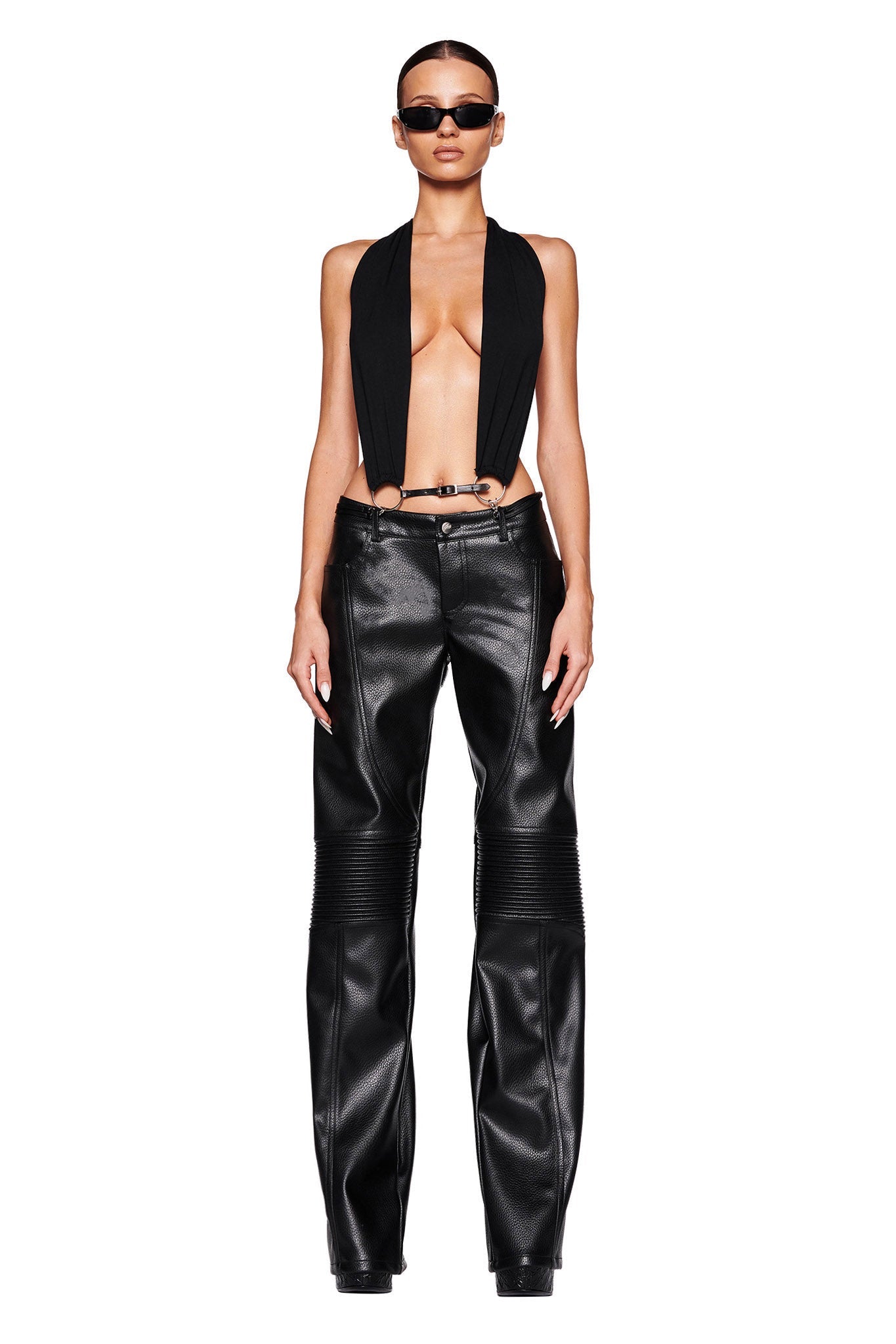A person modeling a stylish I.AM.GIA outfit, featuring the MIKAYLA TOP - BLACK, which is a black sleeveless open-front top, paired with black high-waisted leather pants. They are also wearing dark sunglasses and have their hair pulled back, standing against a plain white background.