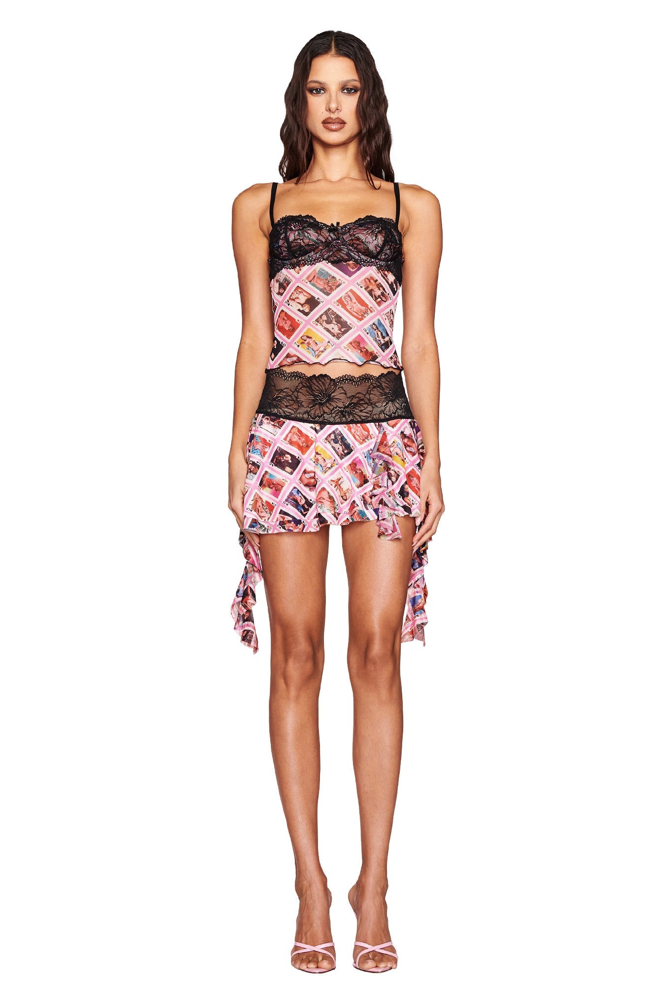 Against a white backdrop, the woman poses in an I.AM.GIA SILO MINI SKIRT - MULTI : GIA FACE CARD featuring colorful patterns and black lace trim. Her strappy high-heeled sandals complement the asymmetrical hem, while her hair falls in loose waves.