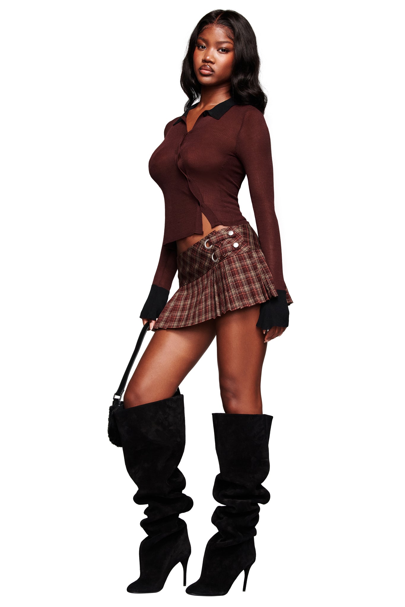 A woman poses in the I.AM.GIA DAVINA TOP - BROWN, a brown plaid pleated mini skirt with lace-up detail, black slouchy knee-high heeled boots, and holds a black handbag.