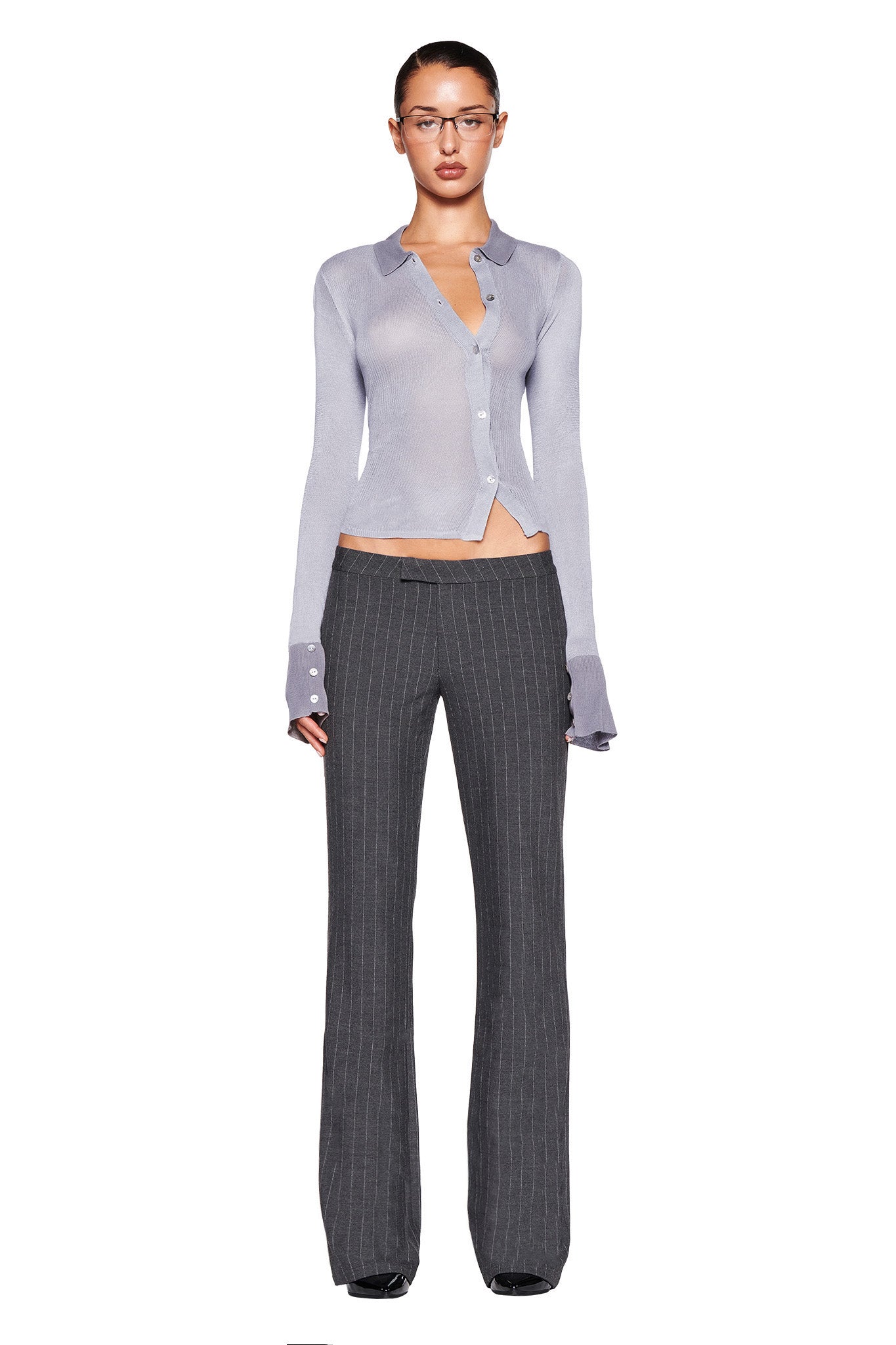 A person stands against a white background wearing the DAVINA TOP - GREY by I.AM.GIA, made of light gray, semi-sheer, high-stretch fabric with thumbholes, paired with dark gray pinstripe pants. They have glasses and their hair is slicked back.