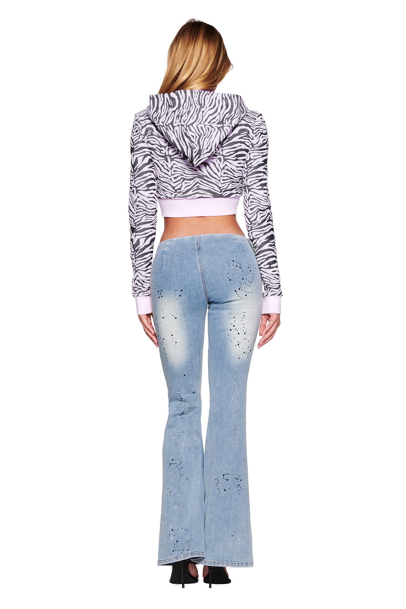 A woman with long blonde hair is facing away, wearing the I.AM.GIA SOLIS HOODIE - PINK: ZEBRA and light-wash, high-waisted flare jeans with small decorative holes. She is standing in high-heeled black shoes, with her arms relaxed at her sides.