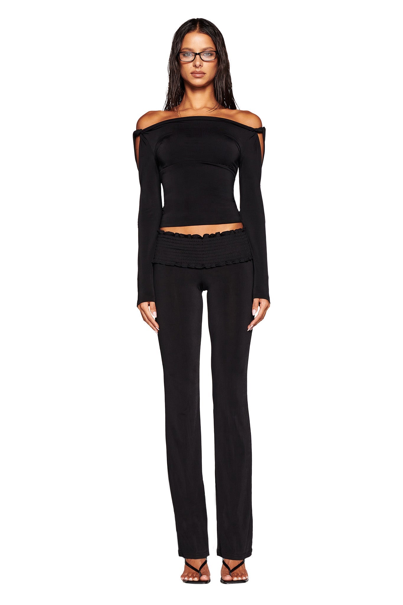 A woman with long dark hair and glasses wears a chic off-the-shoulder black LORELAI TOP from I.AM.GIA, featuring long sleeves, paired with black flared pants. She stands against a white background looking forward, her arms relaxed at her sides and feet in black strappy heels.