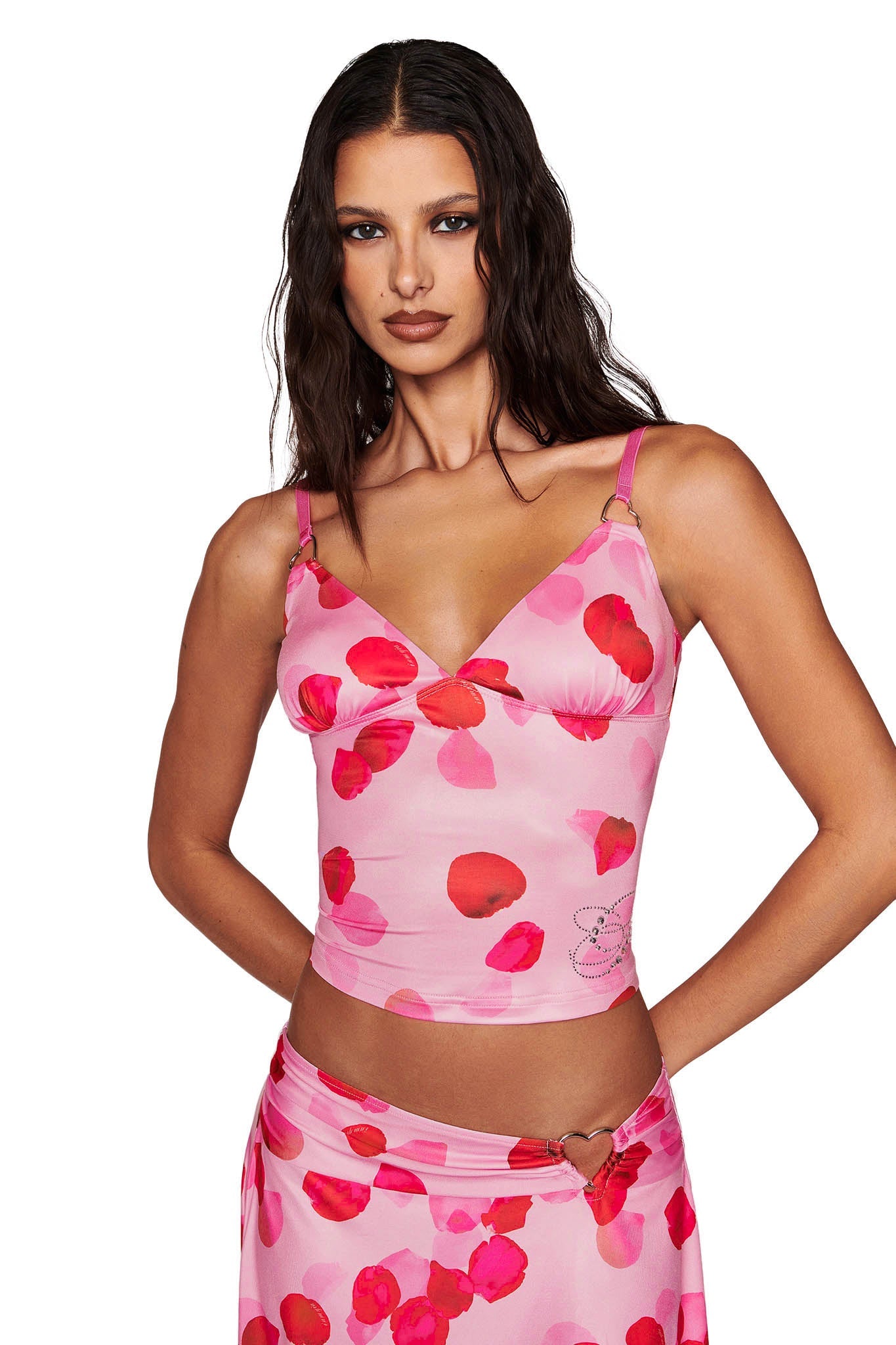A woman poses with her hands on her hips wearing the I.AM.GIA ODETTA TOP - PINK: PRETTY PETAL, adorned with a floral pattern and custom diamantes. She pairs it with a matching skirt, and her long, wavy hair drapes elegantly over her shoulders.