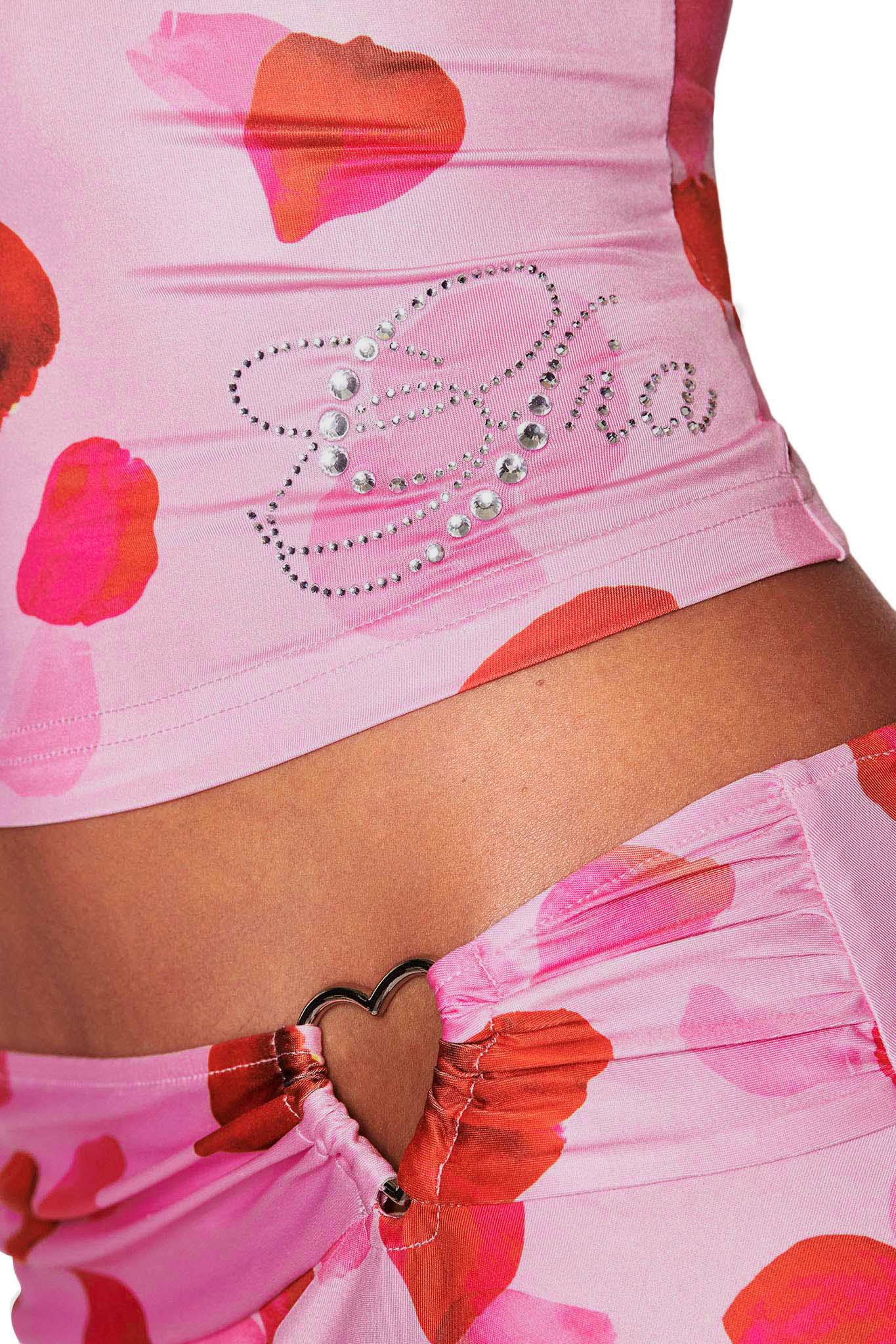 Close-up of a person in the I.AM.GIA ODETTA TOP - PINK : PRETTY PETAL, featuring "Cha" in custom diamantes, with bottoms that have a heart-shaped metal detail on the waistband.