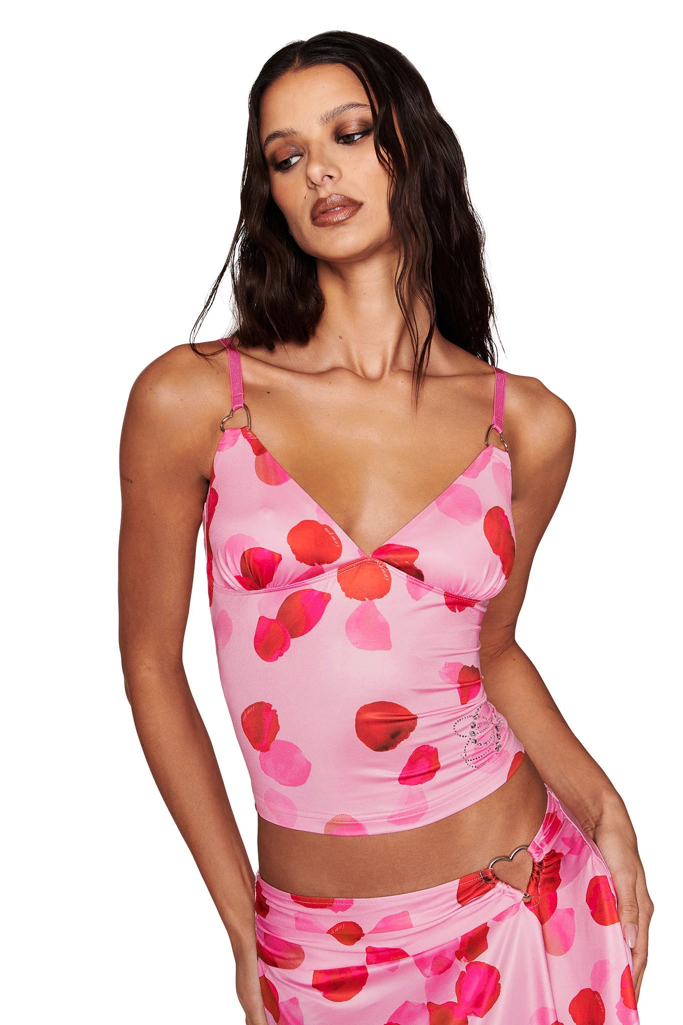 A person with long dark hair is wearing the I.AM.GIA ODETTA TOP - PINK : PRETTY PETAL, adorned with red and pink flowers. The matching skirt features custom diamantes and a heart-shaped detail on a crisp white background.
