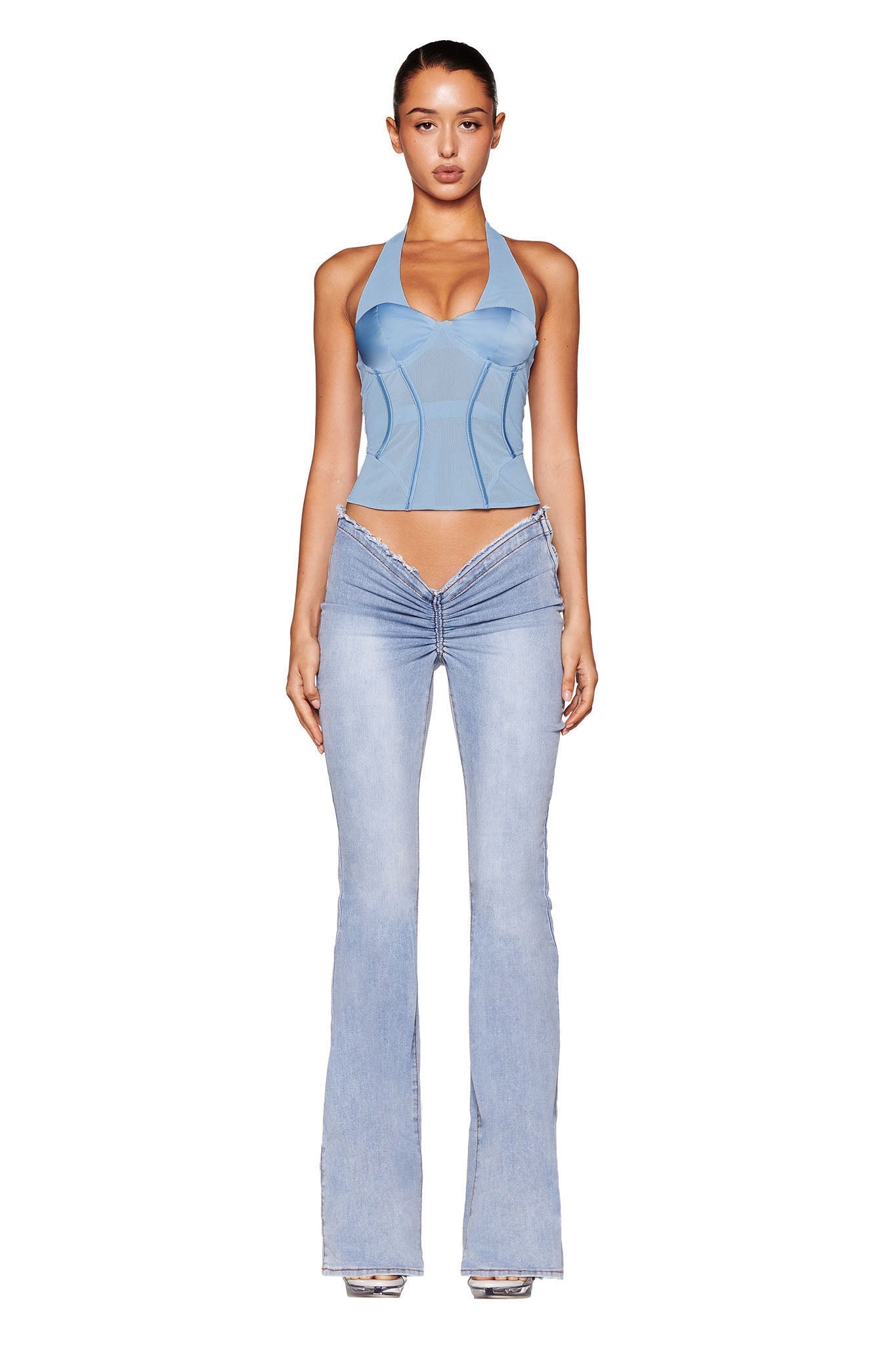 A woman stands against a white background wearing the LOLA Corset Top in Baby Blue by I.AM.GIA, paired with light blue, low-rise flared jeans. She also wears clear heeled shoes. Her hair is pulled back, and she looks directly at the camera.
