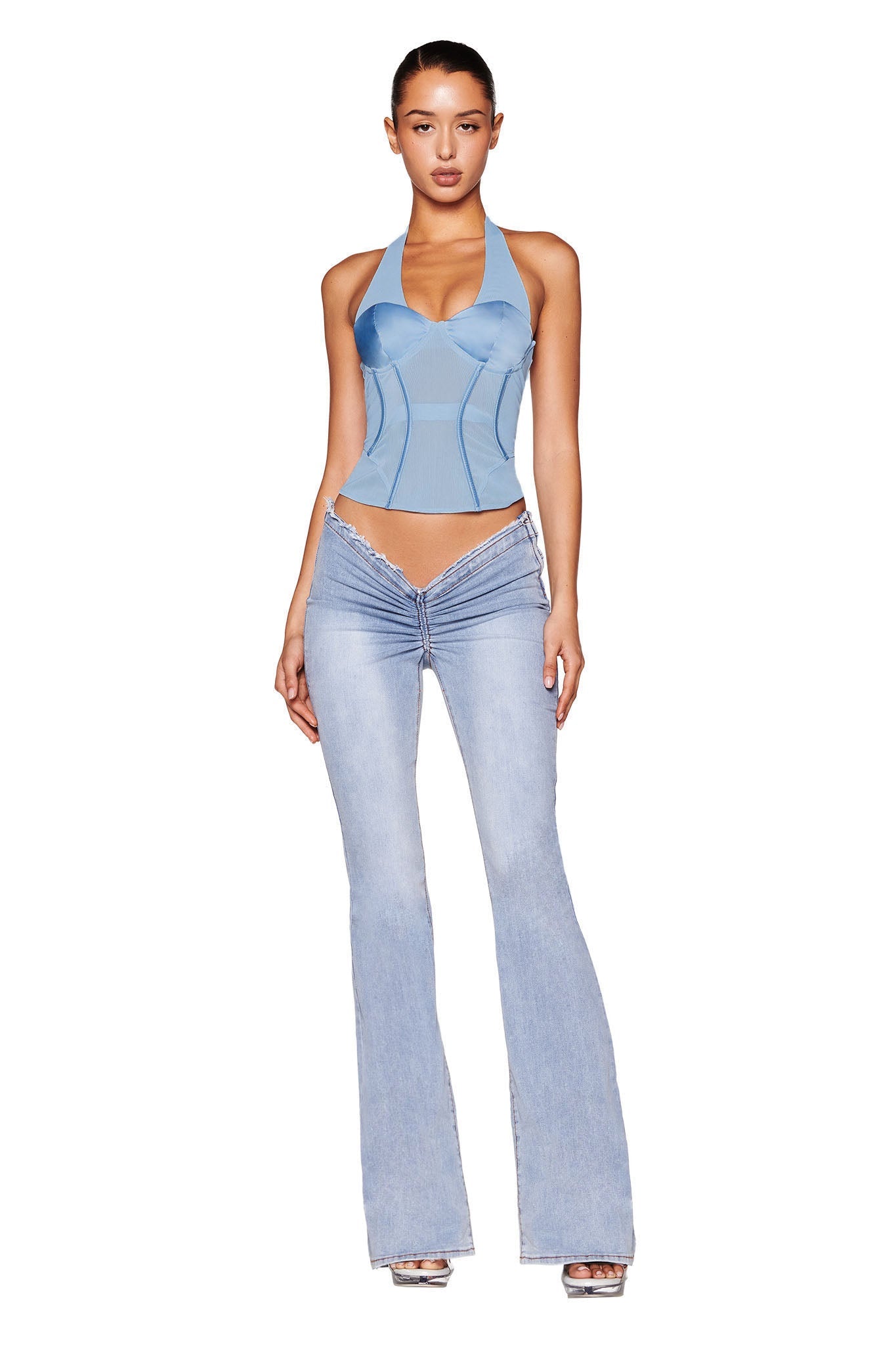 A woman stands wearing the LOLA CORSET TOP in blue, branded by I.AM.GIA, featuring satin bust cups, along with light blue flare jeans and silver high heels. Her long dark hair is tied back as she poses against a plain white background.