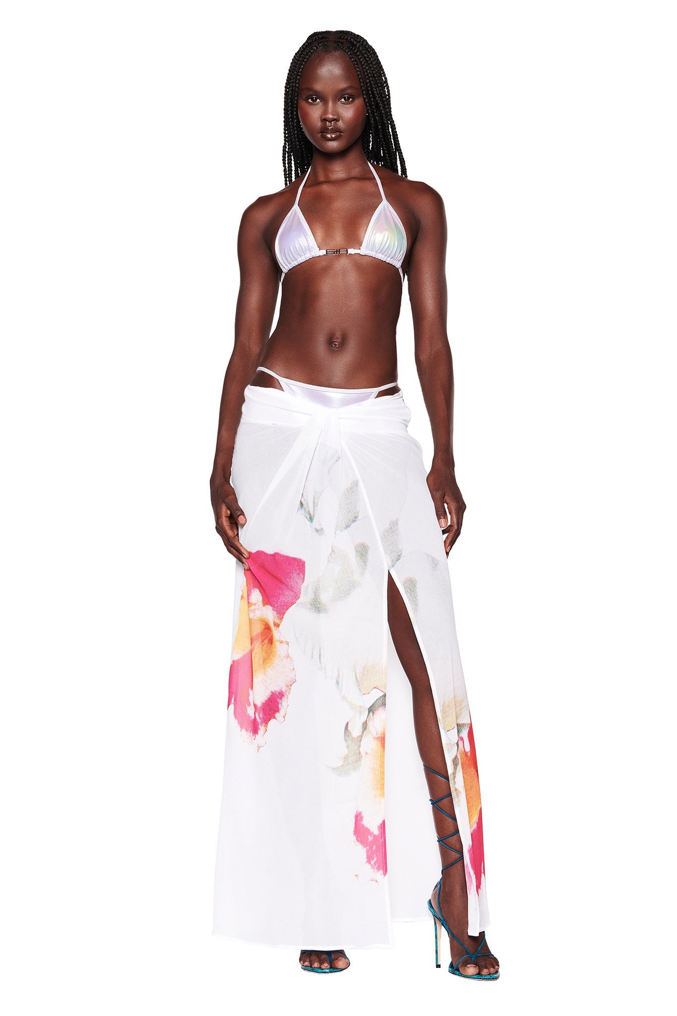 A person with long braided hair stands confidently wearing the I.AM.GIA ALOHI Bikini Top in white pearl, a matching bottom, and a white wrap skirt adorned with colorful abstract patterns. They pair this look with strappy high-heel sandals. The background is plain white.