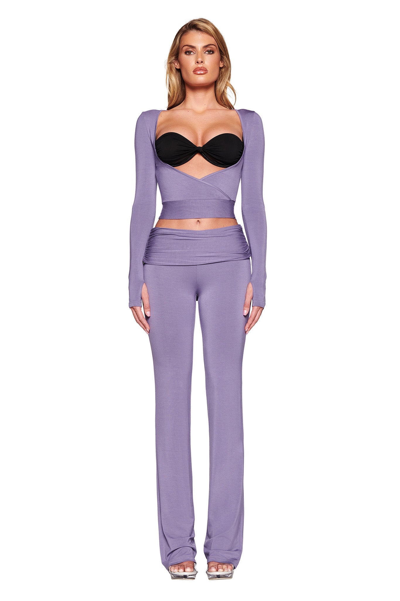 A model is standing against a plain white background, wearing a stylish outfit consisting of a cropped, long-sleeve lavender top with cut-out details and a black bralette underneath, paired with the JANNIS PANT - PURPLE : DUSTY LILAC from I.AM.GIA, featuring a mid-rise waist and flared leg.