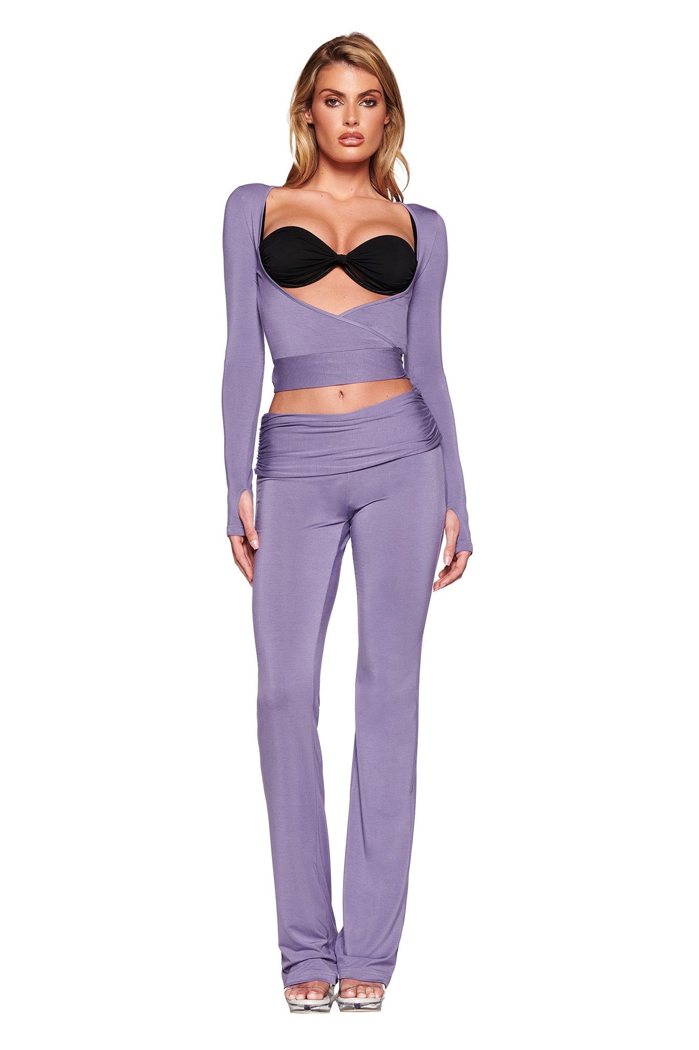 A woman stands against a white backdrop wearing a stylish outfit. She has on the I.AM.GIA STELLA TOP in Dusty Lilac, layered over a black bralette, and matching high-waisted flare pants in soft jersey fabrication. She is also wearing silver high-heeled shoes. Her expression is neutral.
