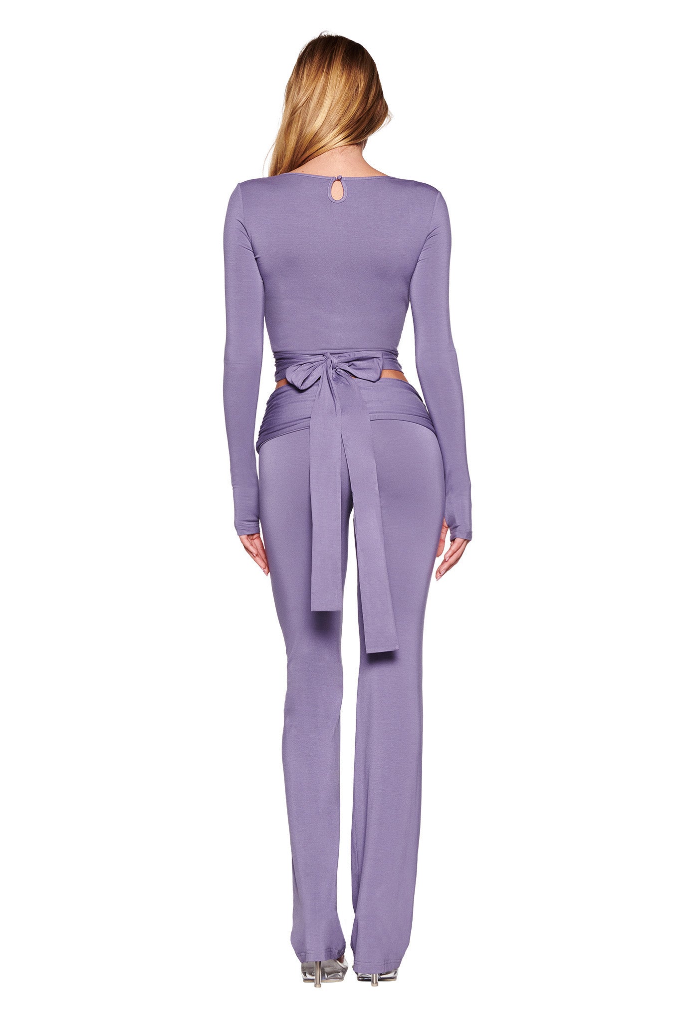 A woman with long blonde hair is seen from the back, wearing a form-fitting, long-sleeved jumpsuit in a dusty lilac color. The bodycon silhouette, known as the STELLA TOP by I.AM.GIA, features a tie at the waist, flared pants, and a small keyhole detail at the top of the back. She is standing on a white background.