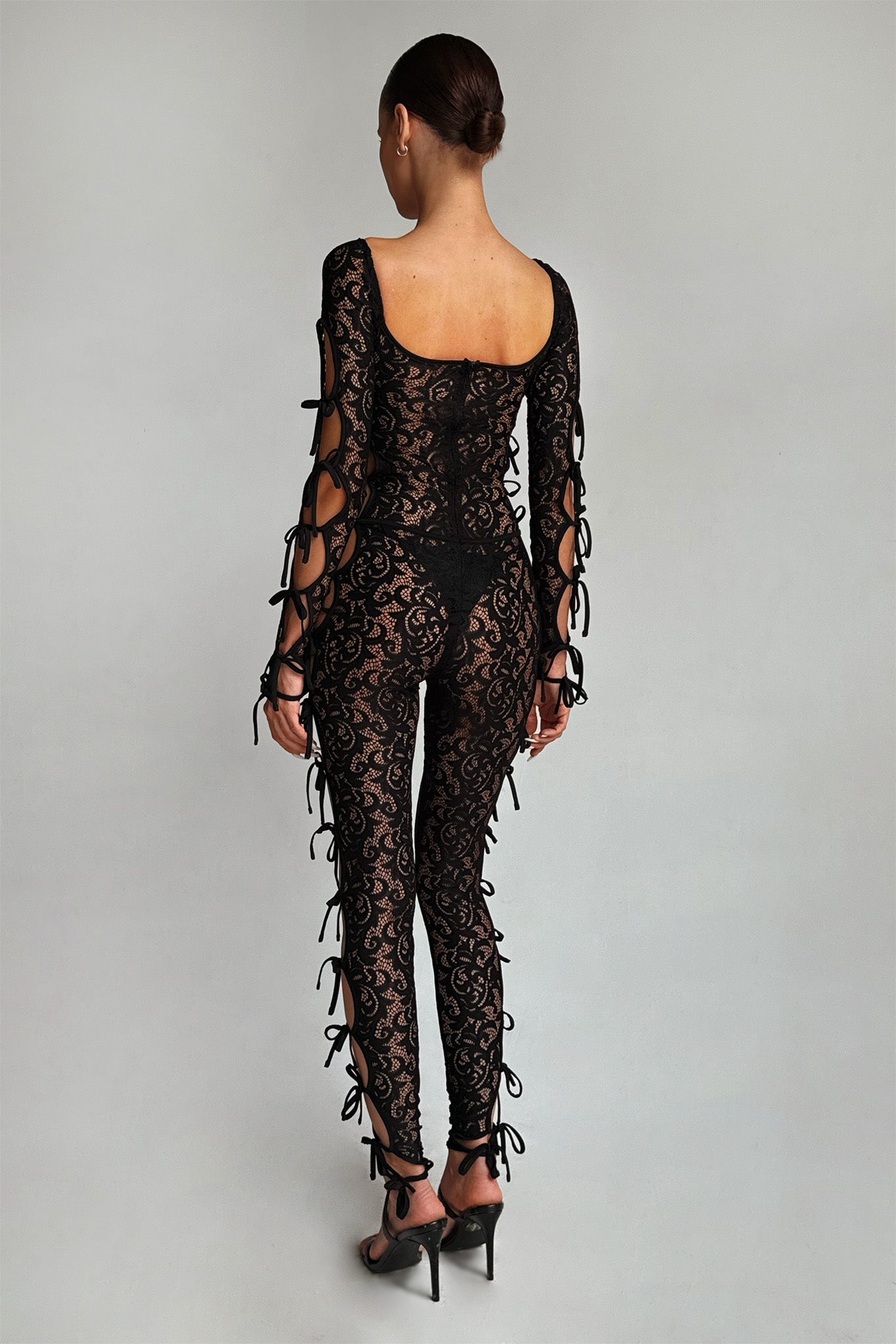 A person stands facing away, wearing a fitted black LYSANDRA CATSUIT - BLACK from the I.AM.GIA brand. This piece features decorative bows along the sides of the arms and legs, a low back, and intricate floral patterns. They are standing against a plain, light grey background.