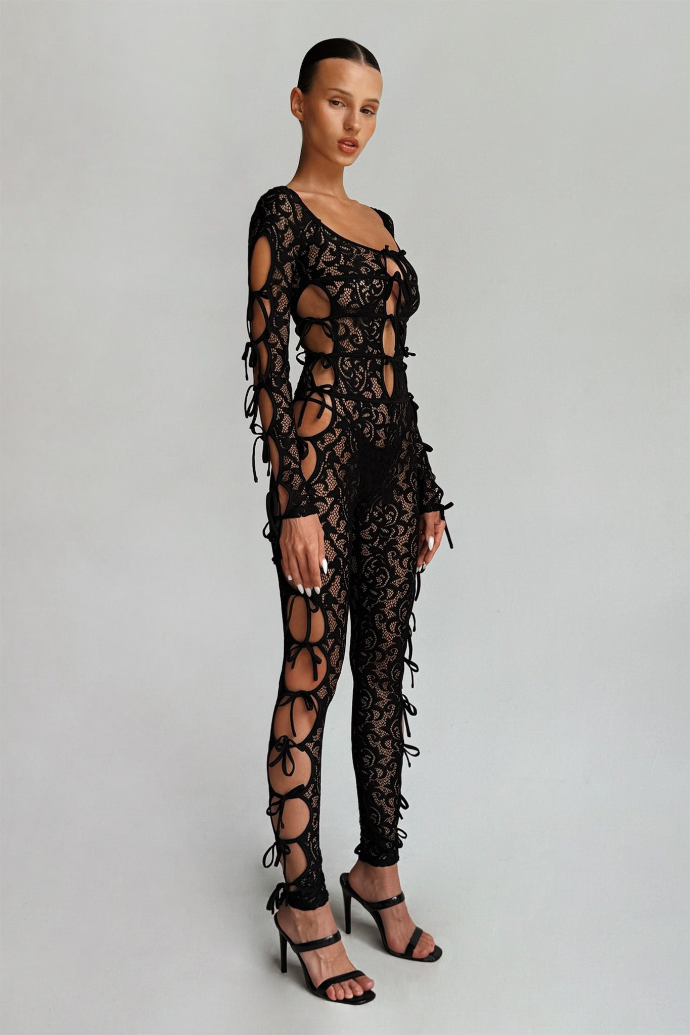 A person stands against a plain background wearing the intricate, all-black Lysandra Catsuit from I.AM.GIA, which features premium lace with cut-out details and thin tie closures. They are also wearing black high-heeled sandals and have their hair slicked back neatly.