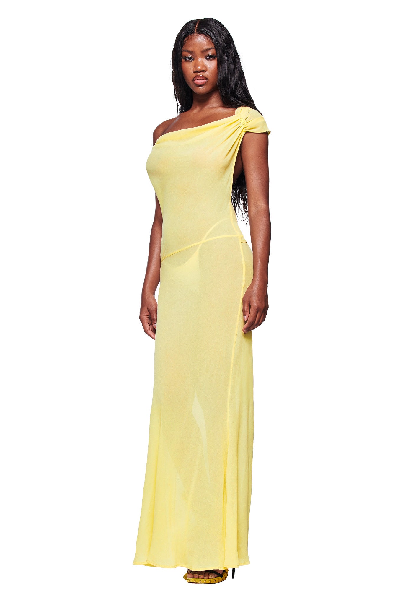 A woman faces forward in the I.AM.GIA KAHLO MAXI DRESS - YELLOW, her long wavy hair flowing over the sheer georgette fabric. She completes the look with yellow heels, all set against a plain white background.